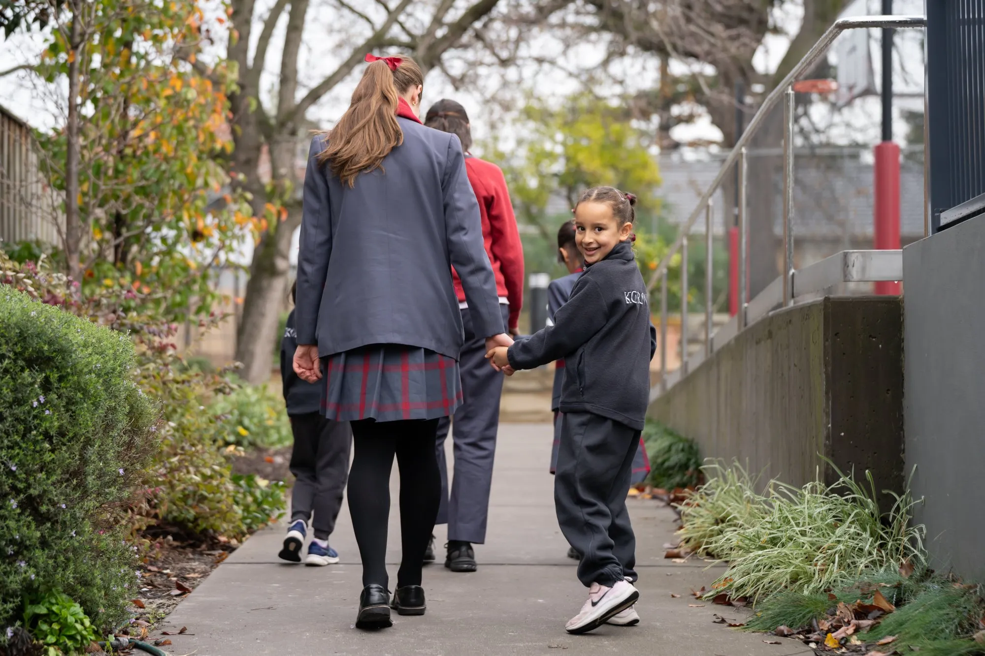 A smiling young girl in a school uniform holding hands with an older student walking on a sidewalk outdoors.