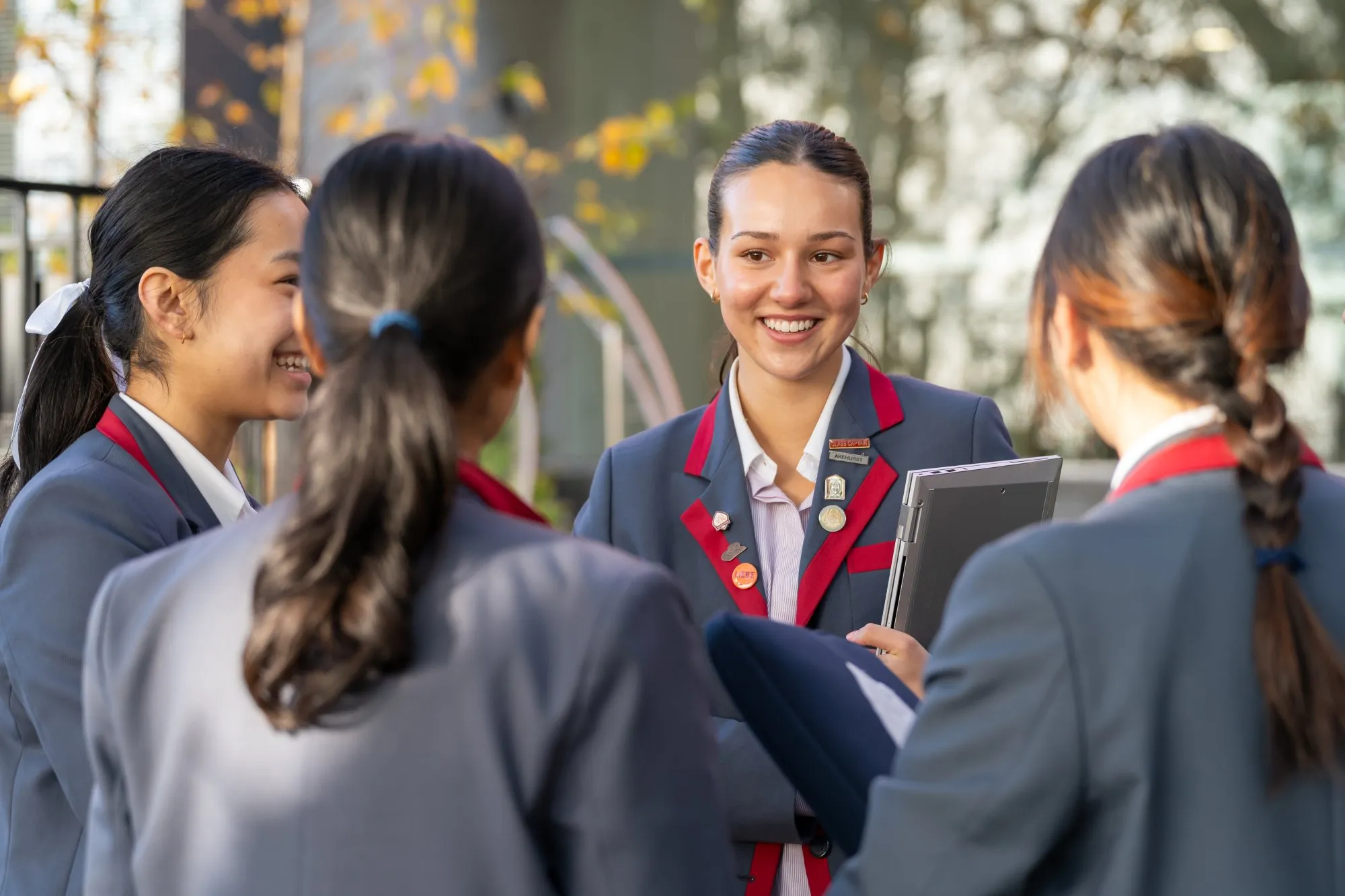 Four female students in gray uniforms with red trim, smiling and talking outdoors, one holding a laptop.