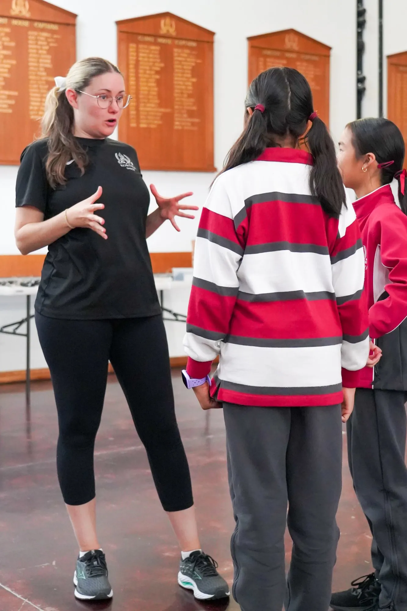 Woman in black athletic clothing speaking and gesturing with hands to two girls in red and white school uniforms inside a hall with wooden plaques on the wall.