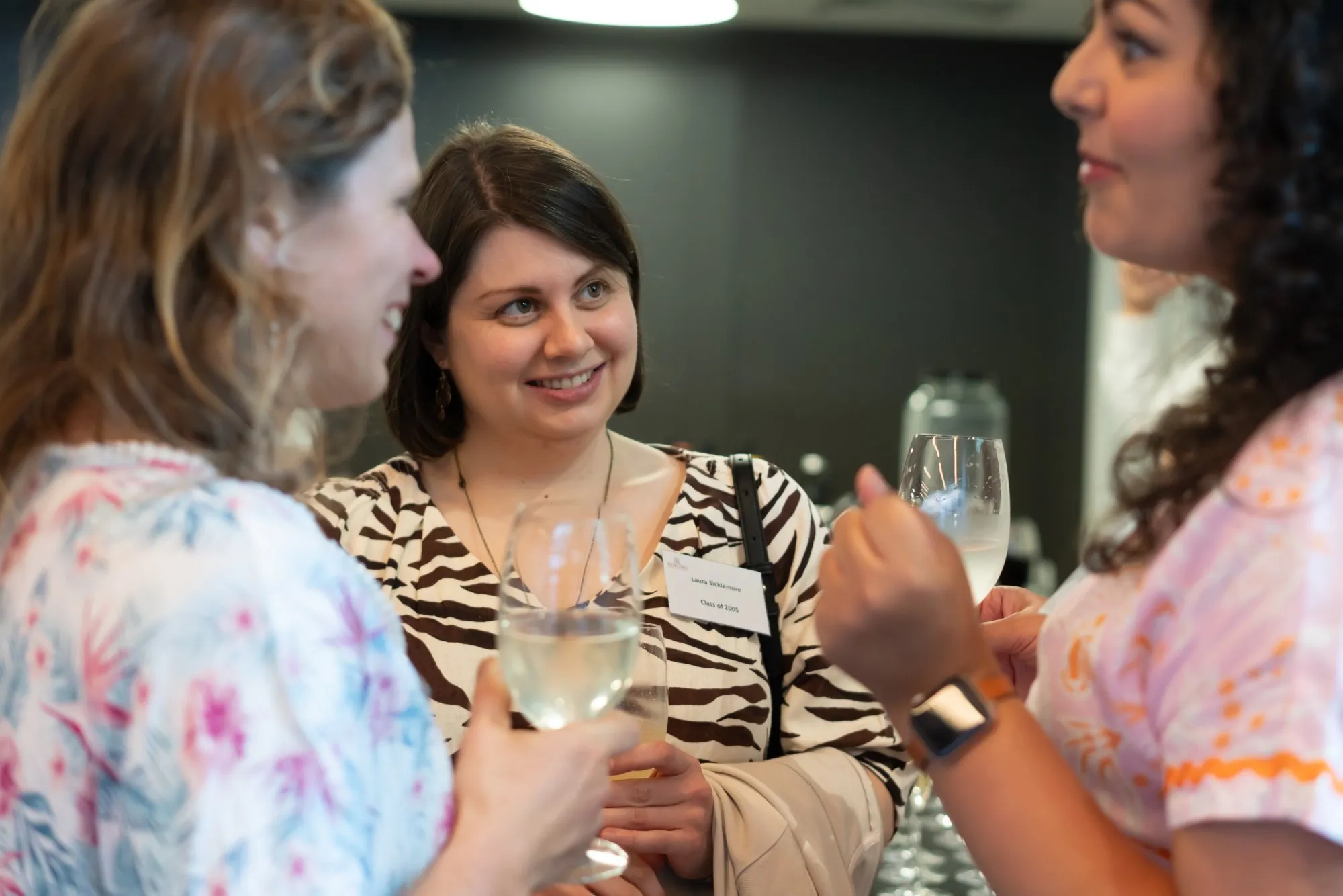 Three women smiling and talking while holding wine glasses at a social event.