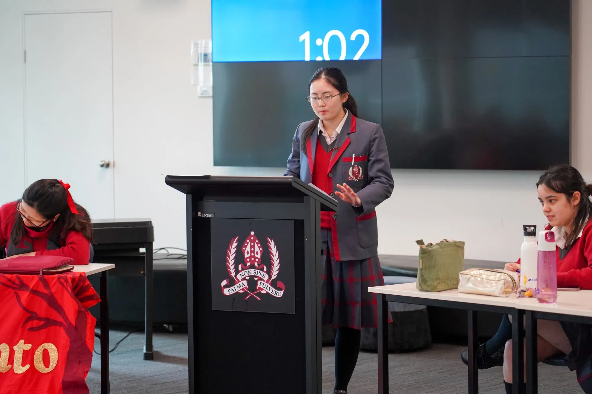 Female student in uniform speaking at a podium in a classroom with two other students seated at tables.