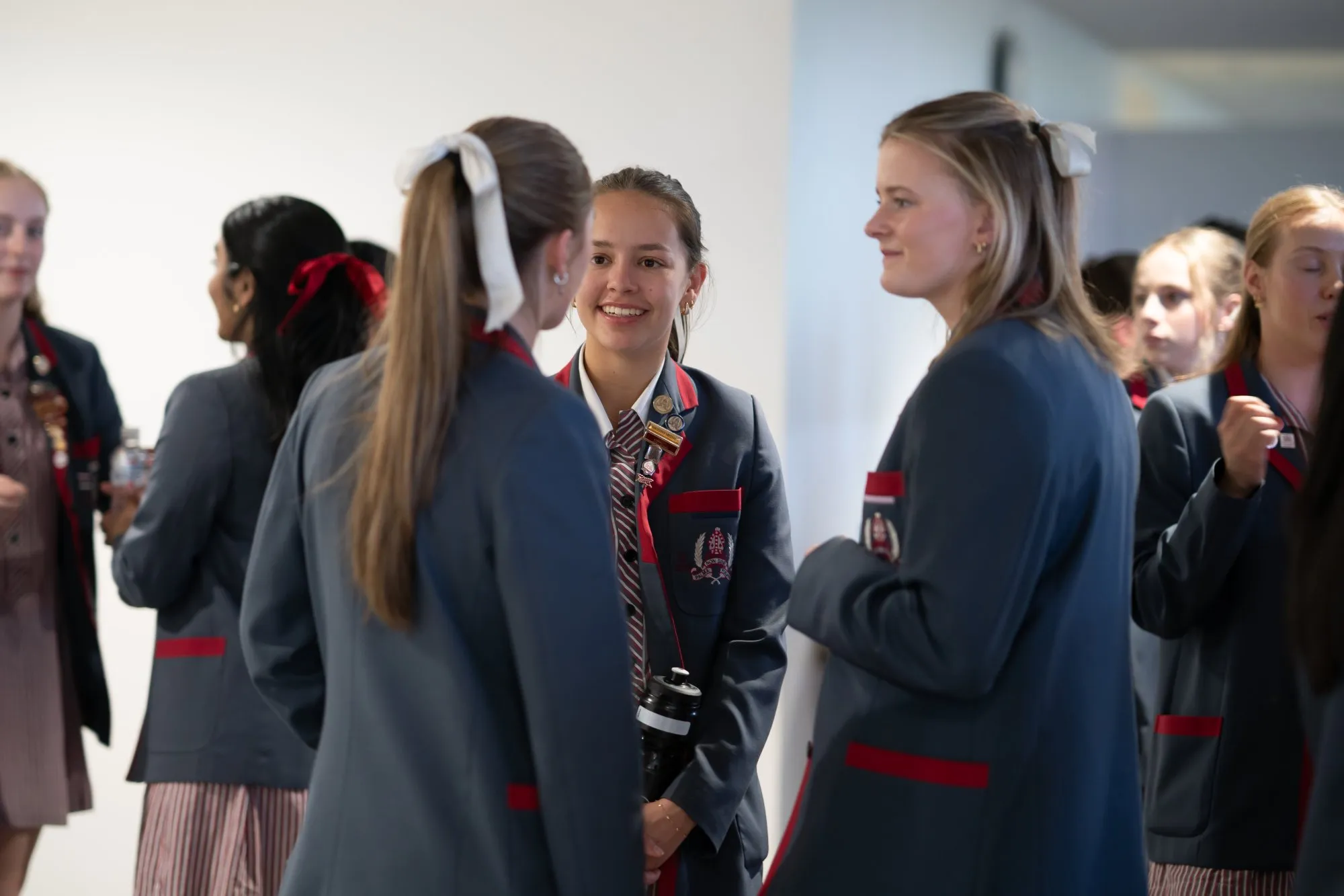 Group of smiling schoolgirls in blue blazers with red trim and striped ties, chatting indoors.