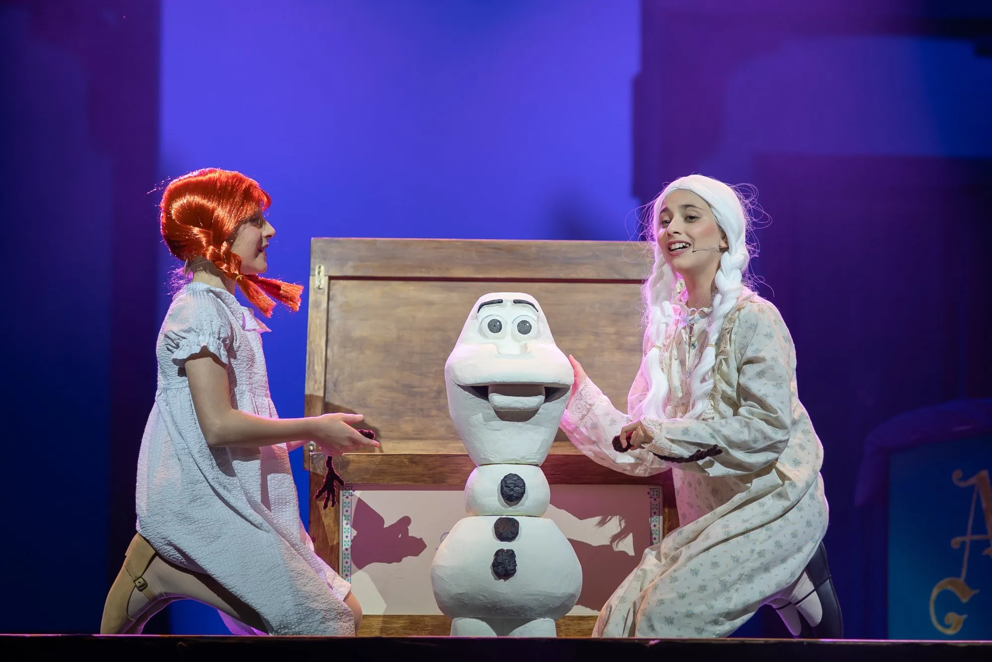 Two performers dressed as children with braided wigs kneel beside a large snowman puppet on stage in a theatrical setting.