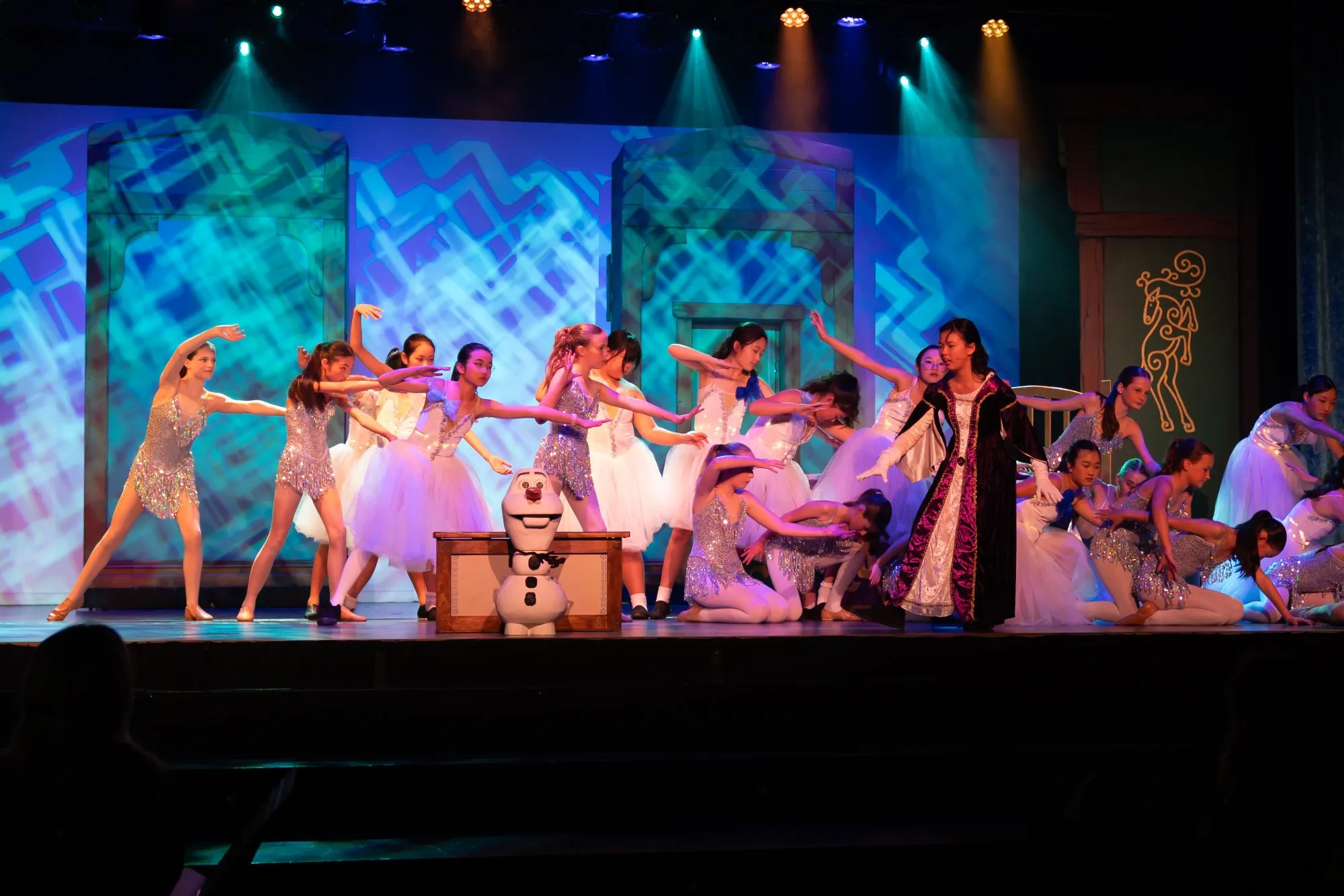 Group of young ballet dancers performing on stage with a snowman prop and colorful stage lighting.