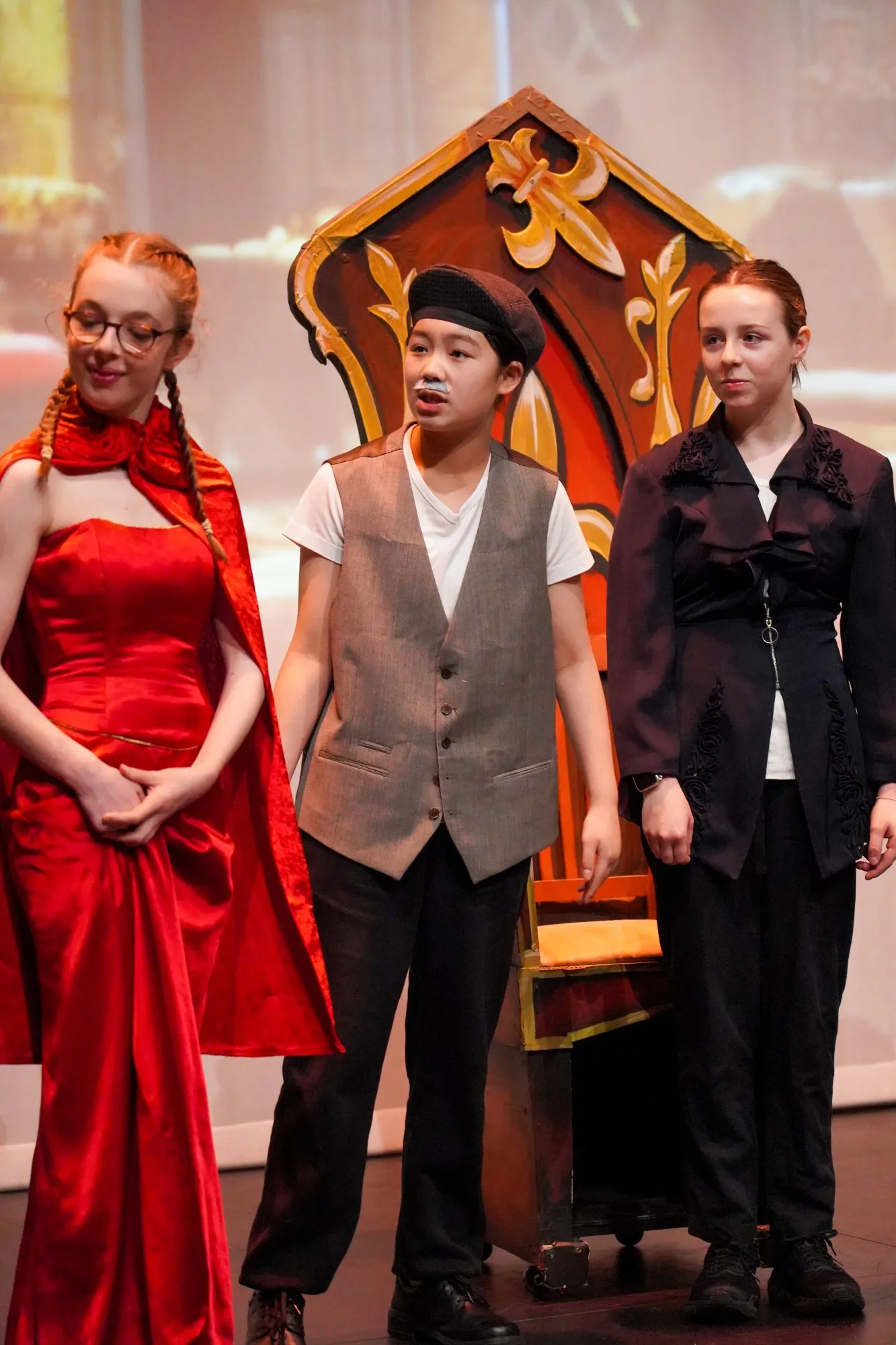 Three young actors on stage with a large ornate chair prop behind them; the girl on the left wears a red dress and cape, the boy in the middle wears a gray vest and cap with a painted mustache, and the girl on the right wears a dark jacket and pants.