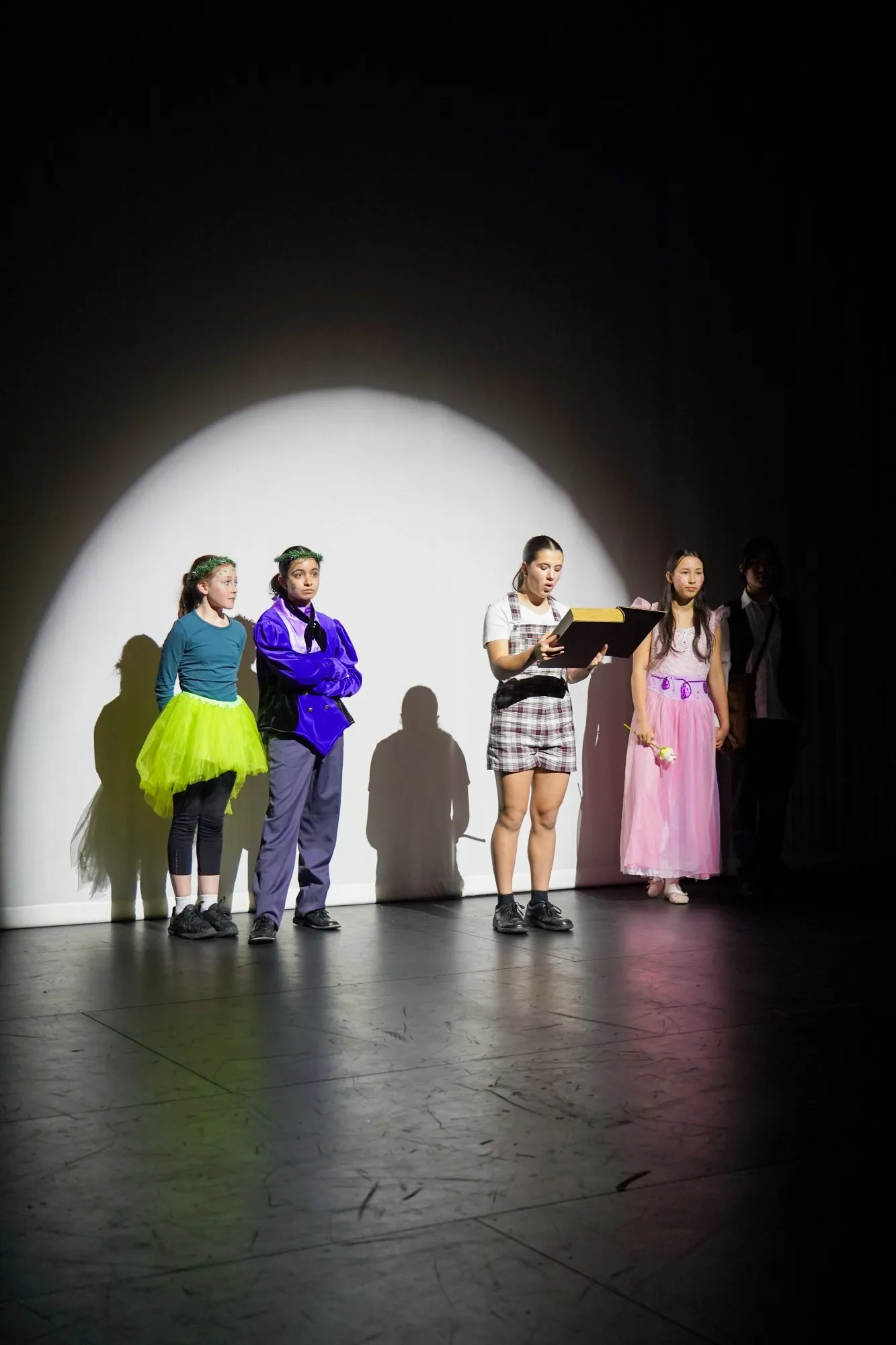 Five children dressed in colorful costumes standing on stage under a spotlight during a theatrical performance.