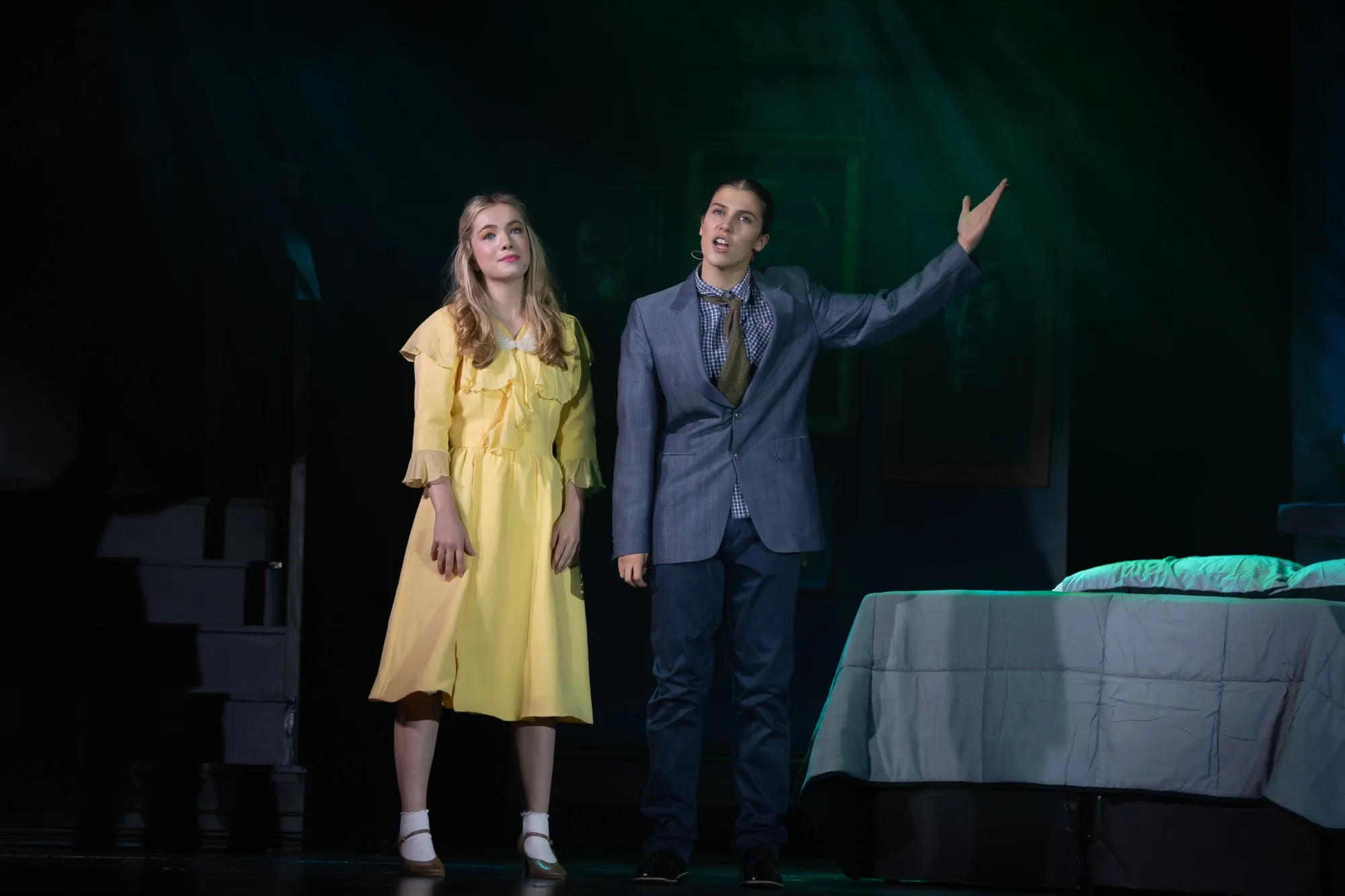A woman in a yellow dress and a man in a suit with a loosened tie perform on stage beside a bed with green lighting.