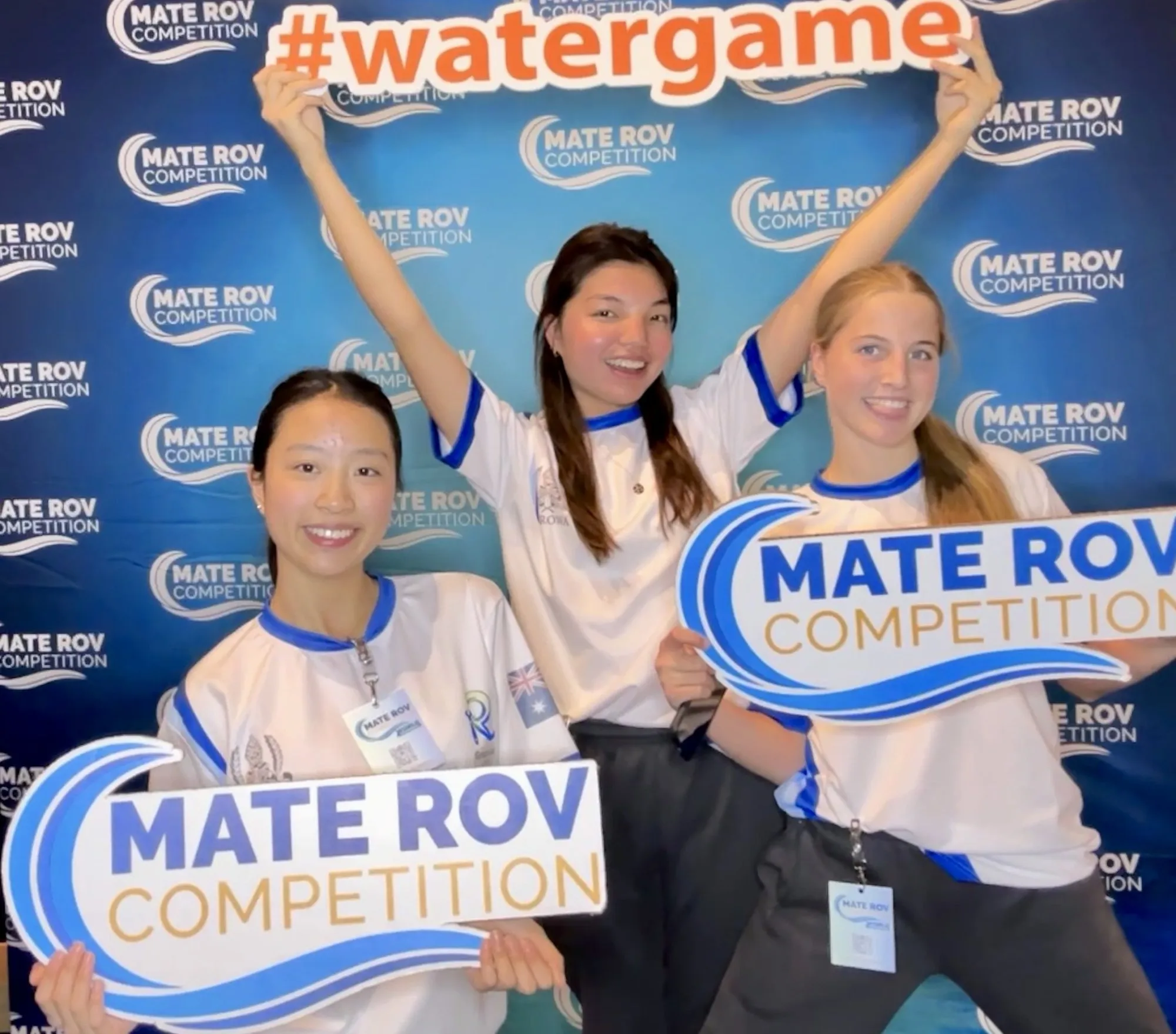 Three young women smiling and holding signs that say 'MATE ROV COMPETITION' and '#watergame' against a blue MATE ROV backdrop.