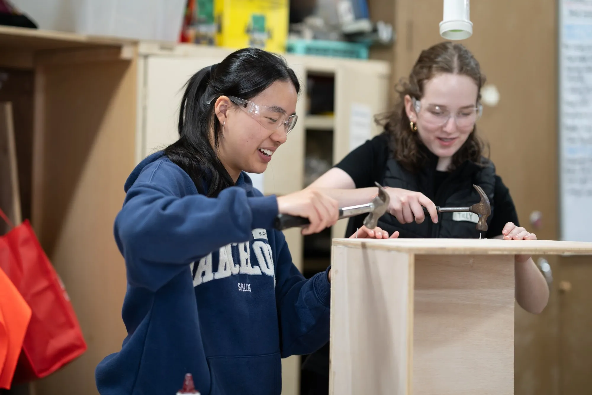 Two young women wearing safety glasses hammering nails into a wooden structure in a workshop.