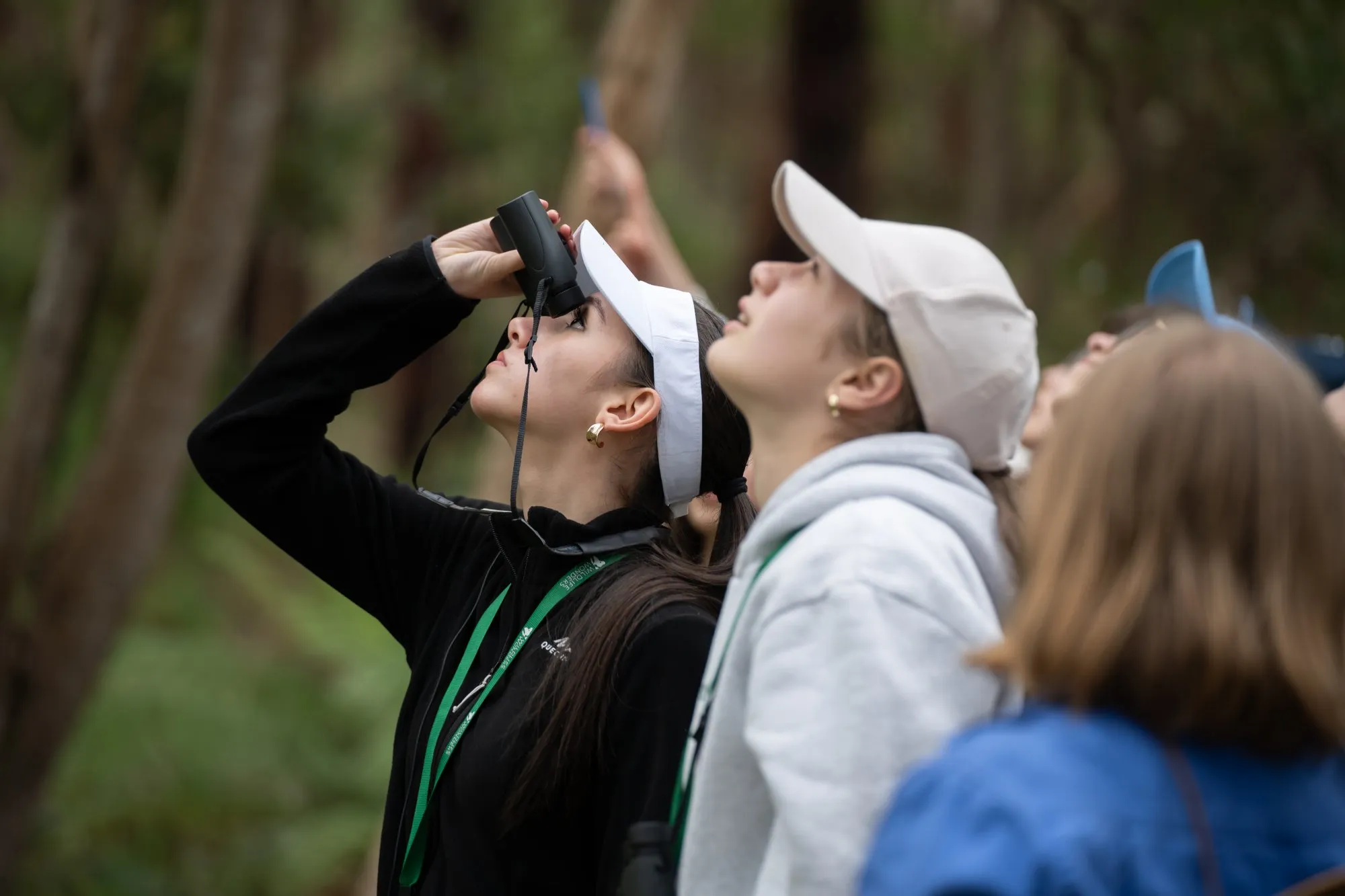 Group of young people outdoors looking up, one woman using binoculars in a wooded area.
