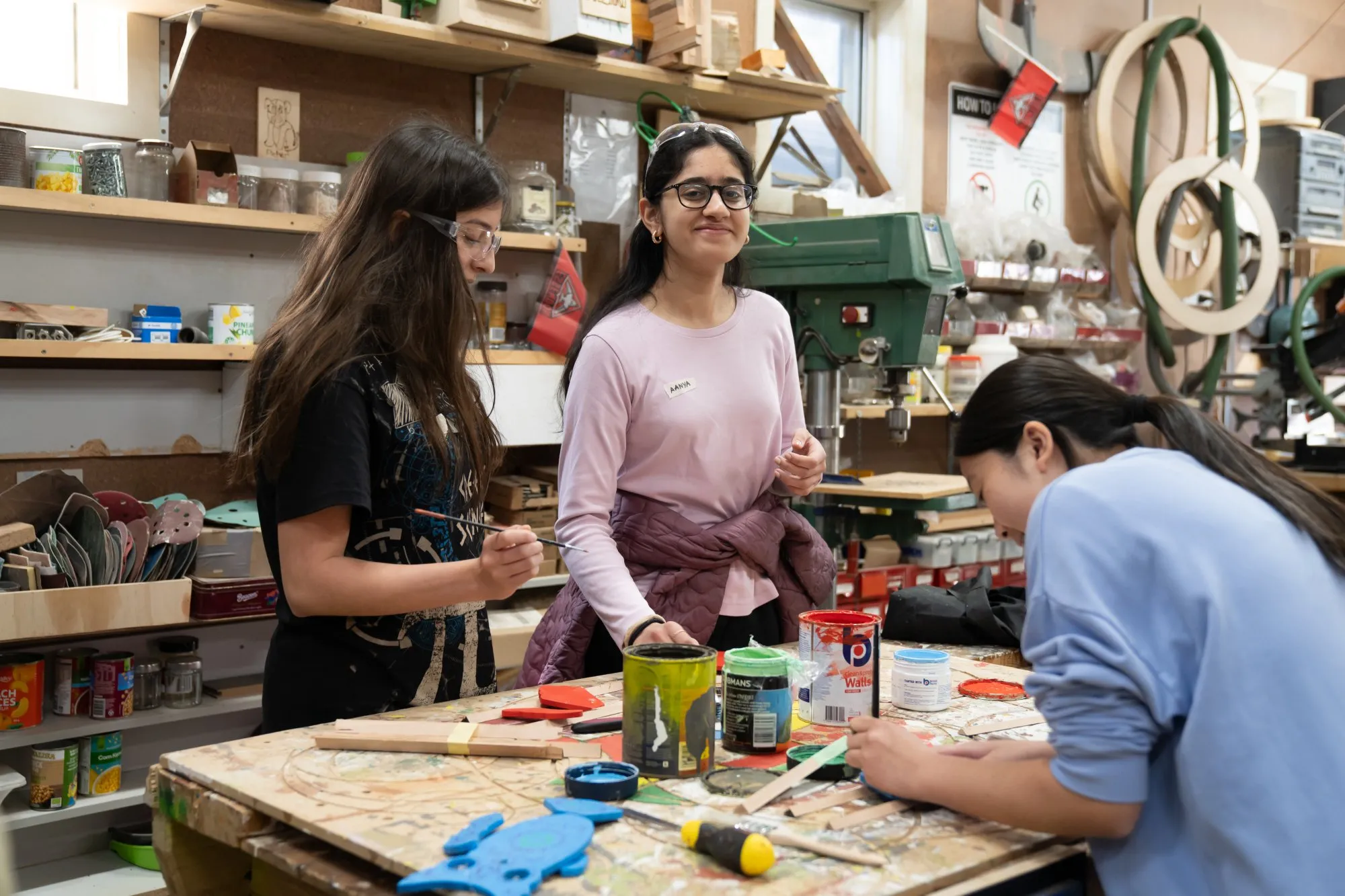 Three young women wearing protective glasses working with paints and craft materials on a wooden table in a workshop.