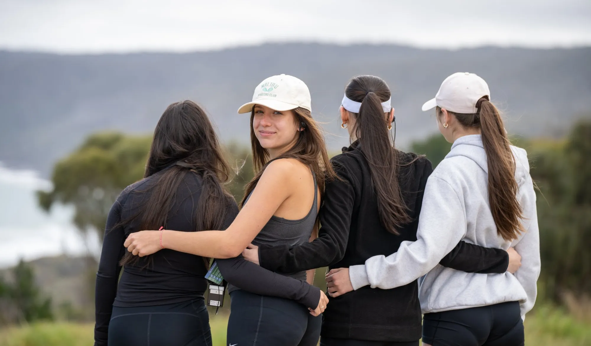Four women with long hair standing side by side outdoors, arms around each other, with two wearing white caps and one looking at the camera and smiling.