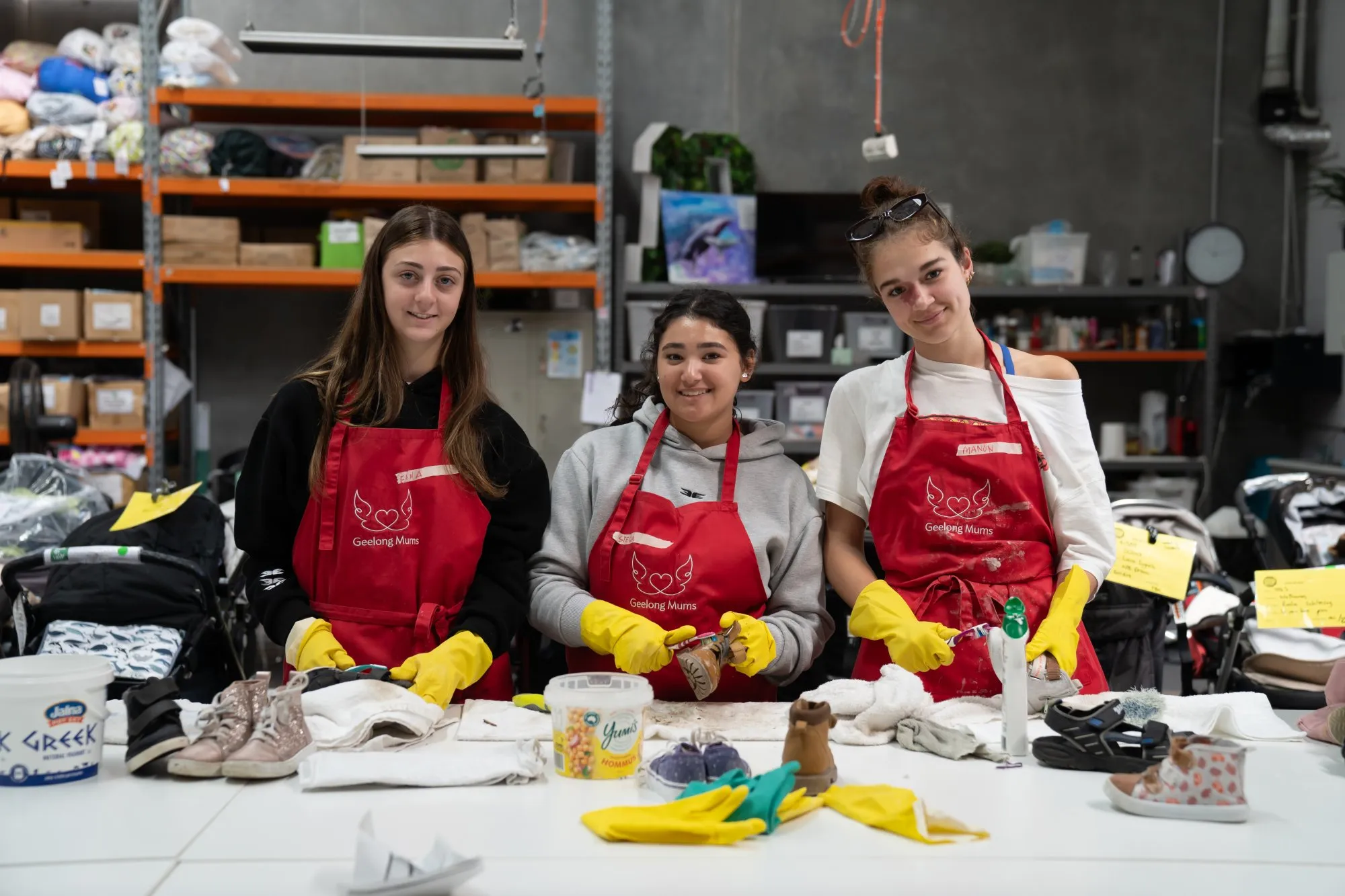 Three young women wearing red Geelong Mums aprons and yellow gloves cleaning baby shoes at a table in a warehouse.