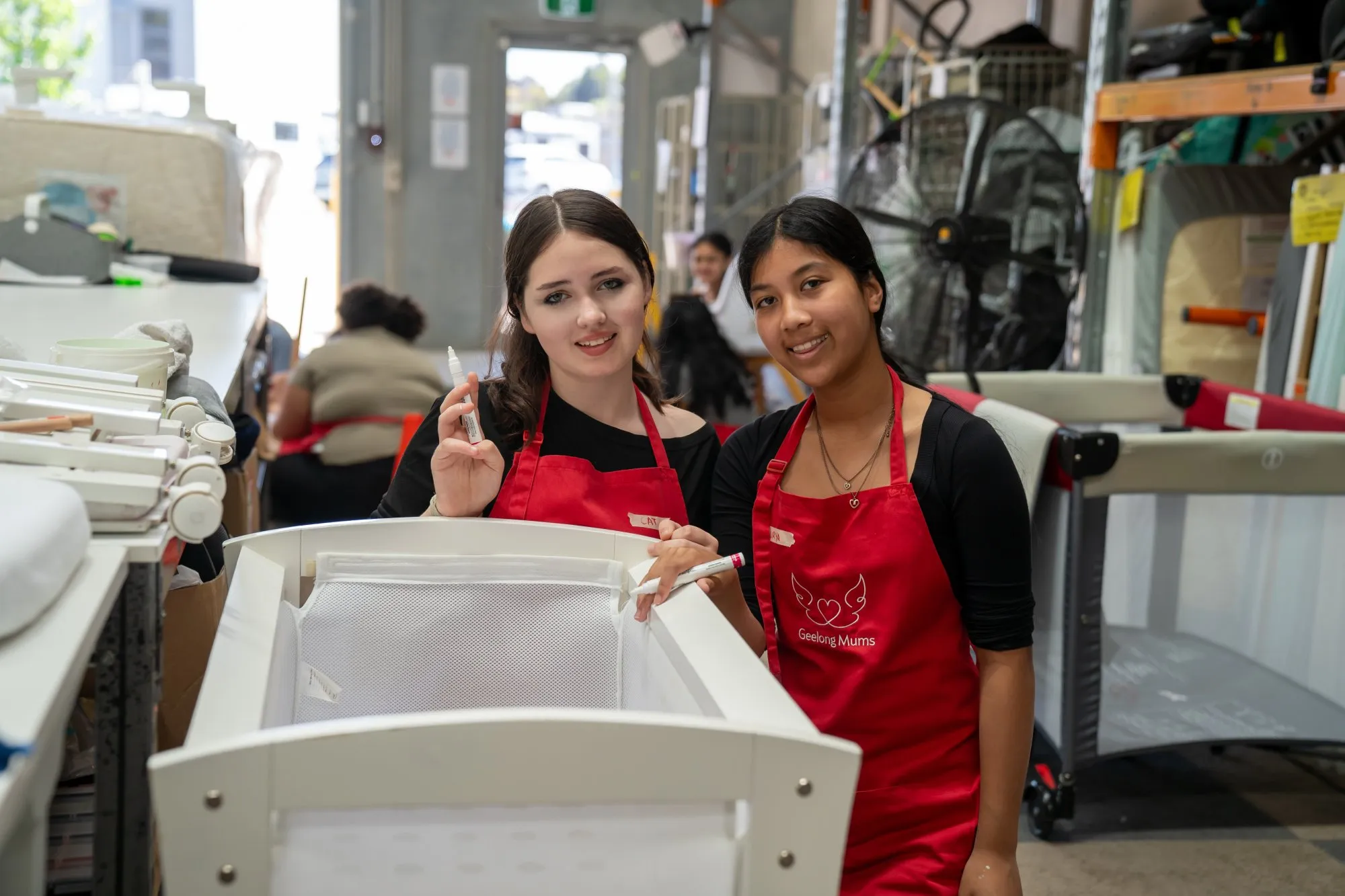 Two women wearing red Geelong Mums aprons standing behind a white baby bassinet in a warehouse setting.