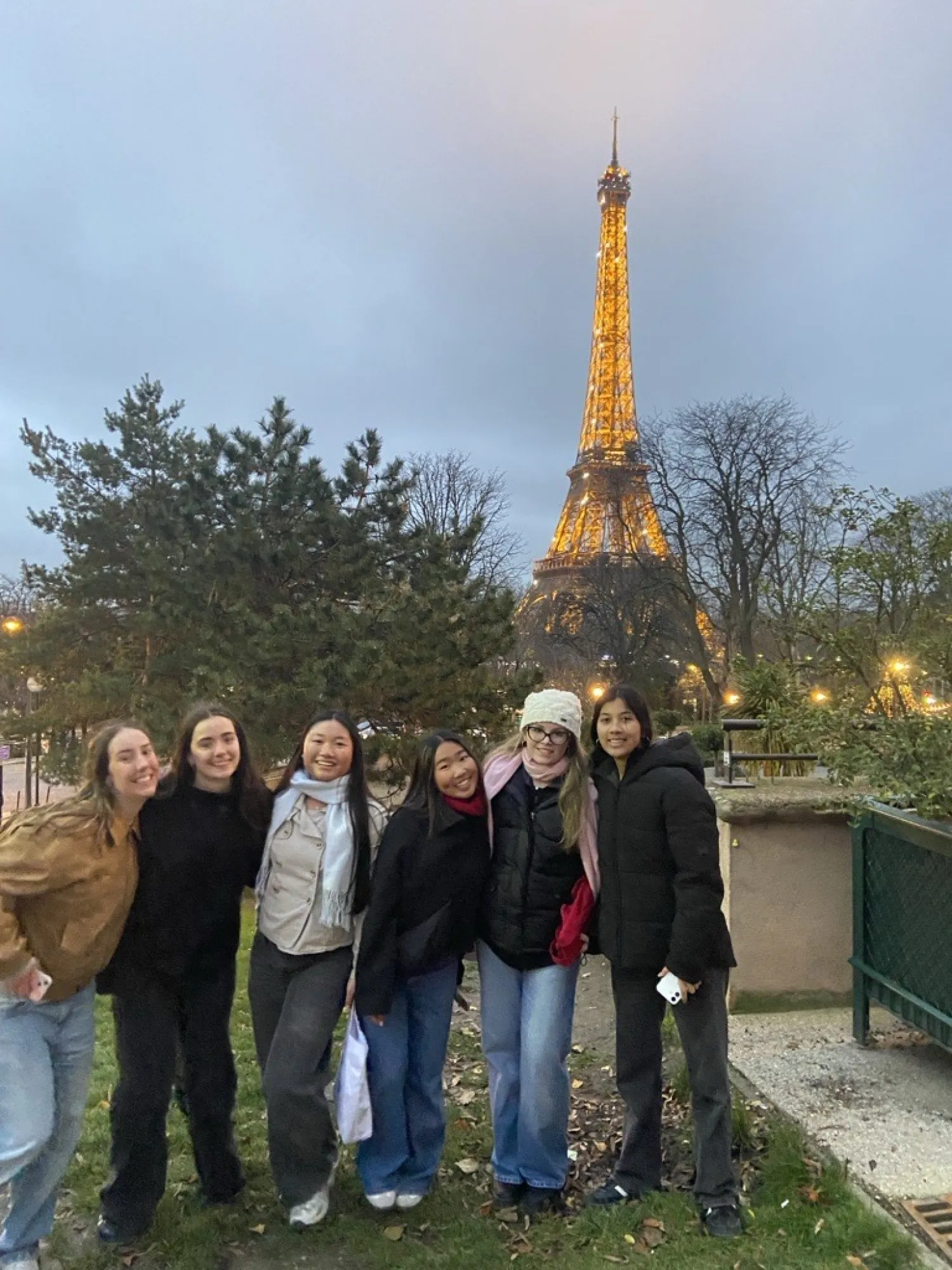 Group of six young women smiling and posing in front of the illuminated Eiffel Tower at dusk.