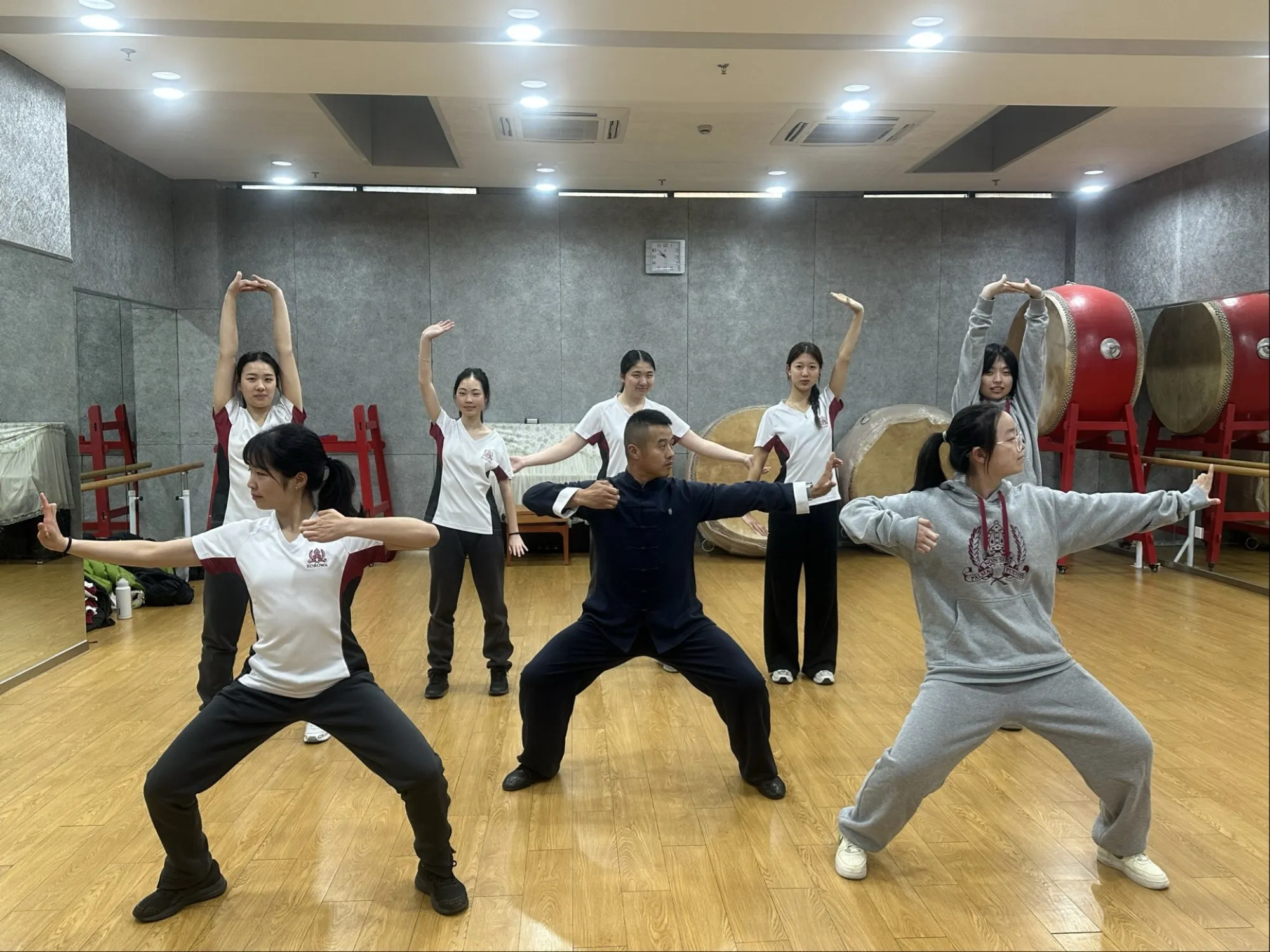 Seven people practicing martial arts poses in a mirrored studio with wooden floor and large drums in the background.