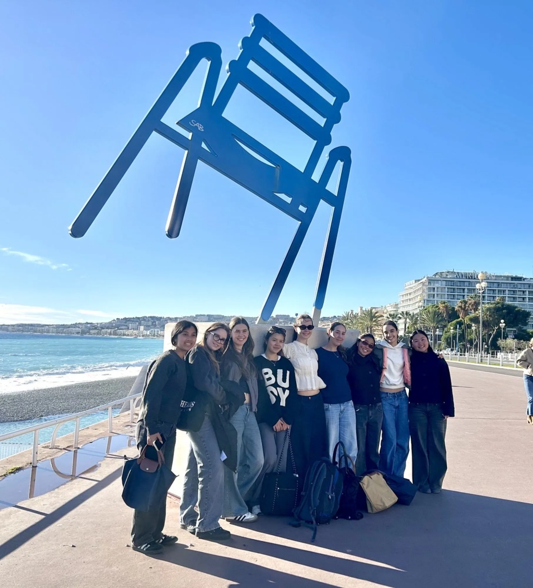 Group of nine young women posing in front of a large blue chair sculpture by the seaside on a sunny day.