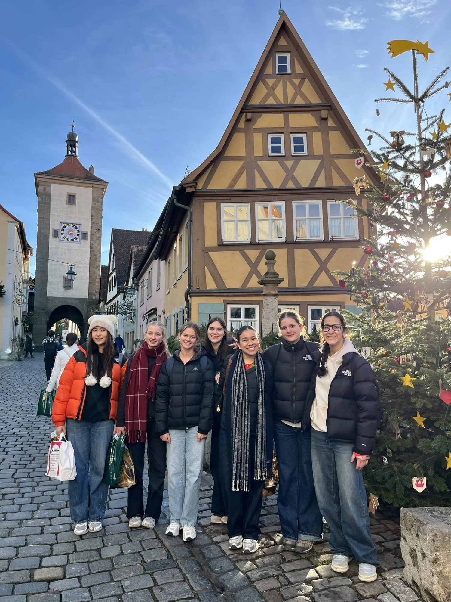 Group of six young women standing on a cobblestone street with traditional half-timbered houses and a clock tower in the background, near a decorated Christmas tree.