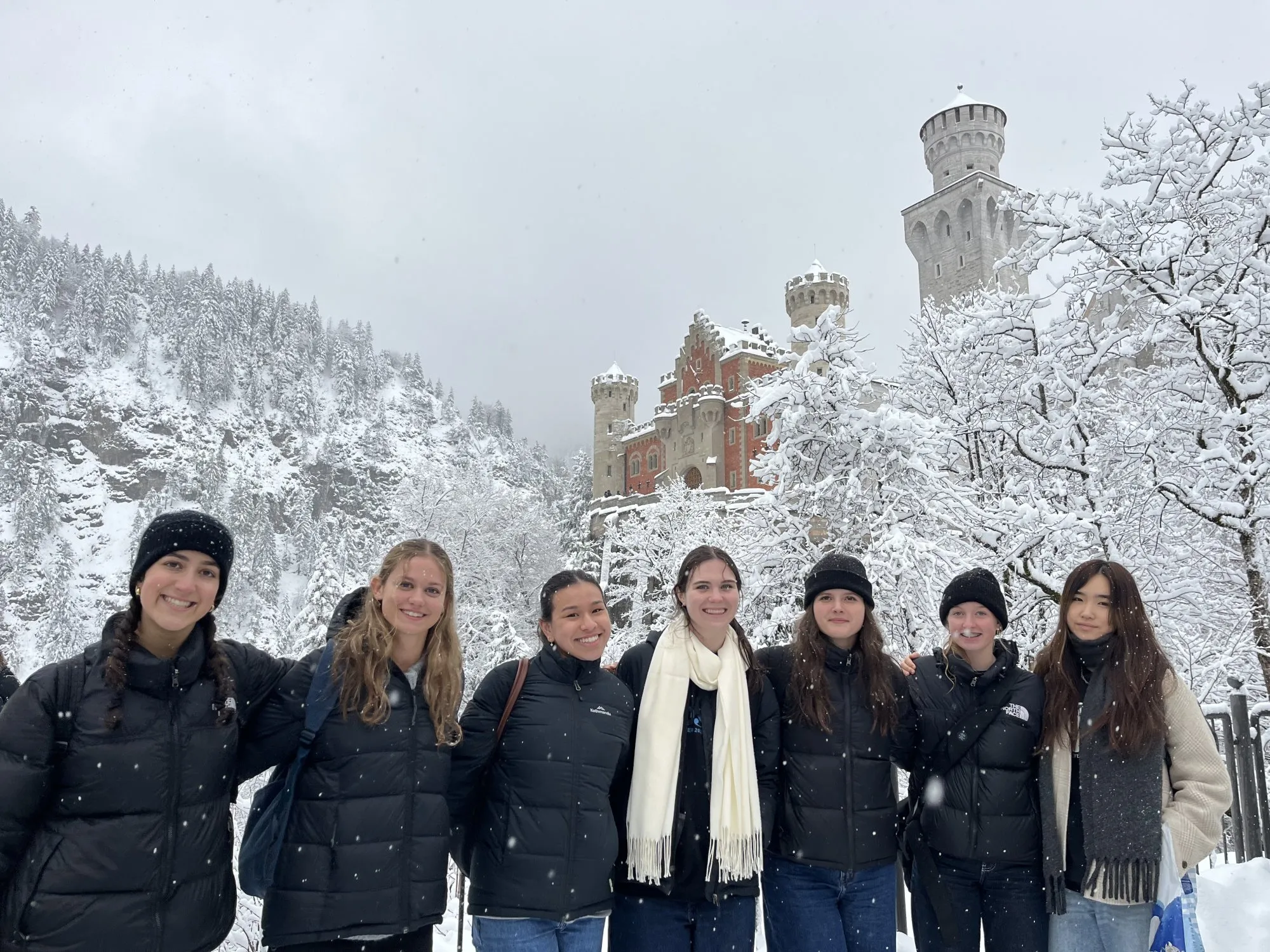 Group of seven young women wearing winter clothing posing in front of a snow-covered castle and trees.