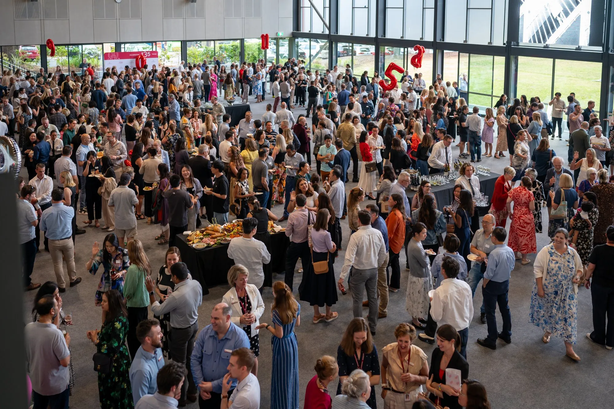Large indoor gathering of professionally dressed people mingling and socializing around tables with food and drinks.