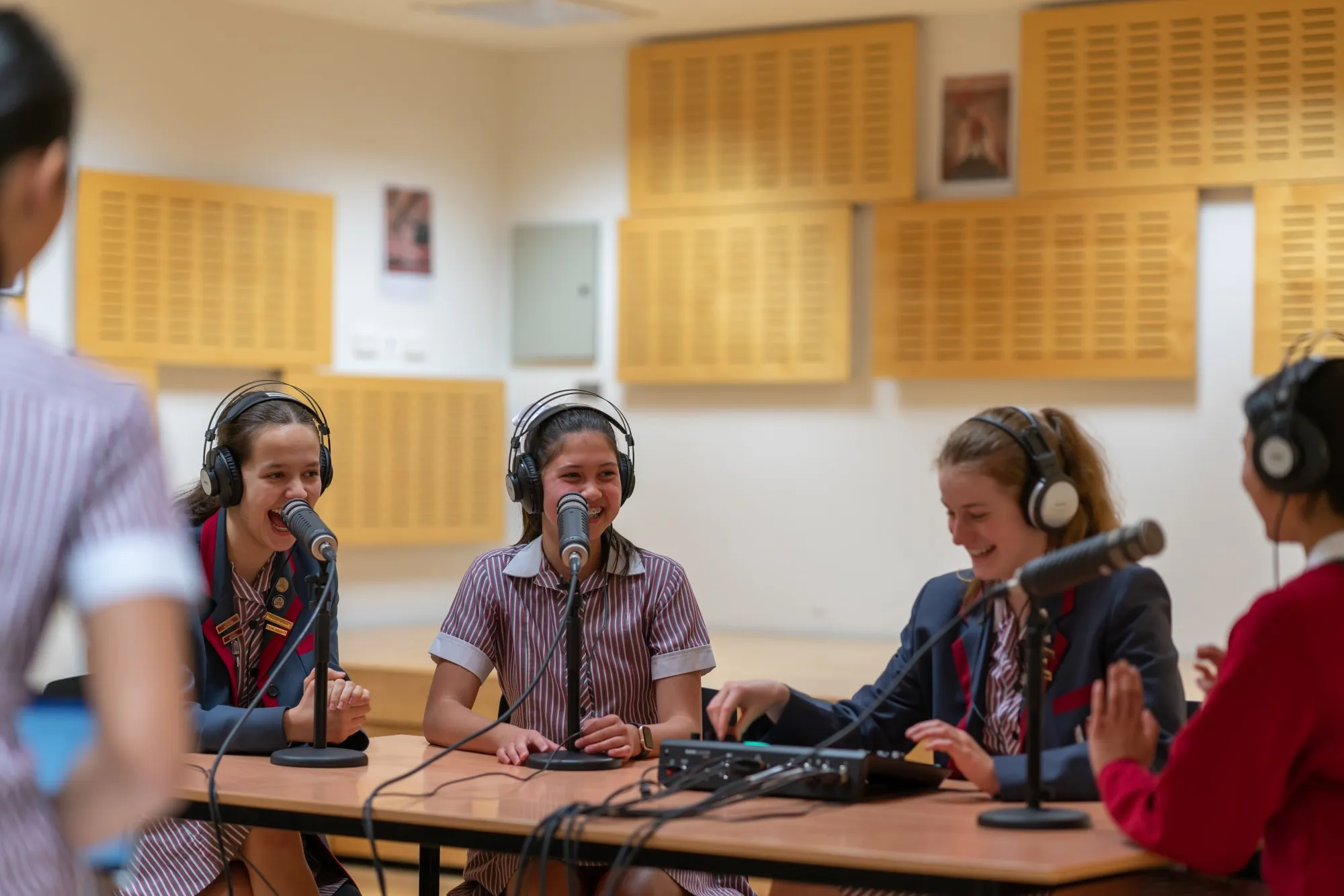 Four teenage girls in school uniforms wearing headphones and speaking into microphones during a recording session.