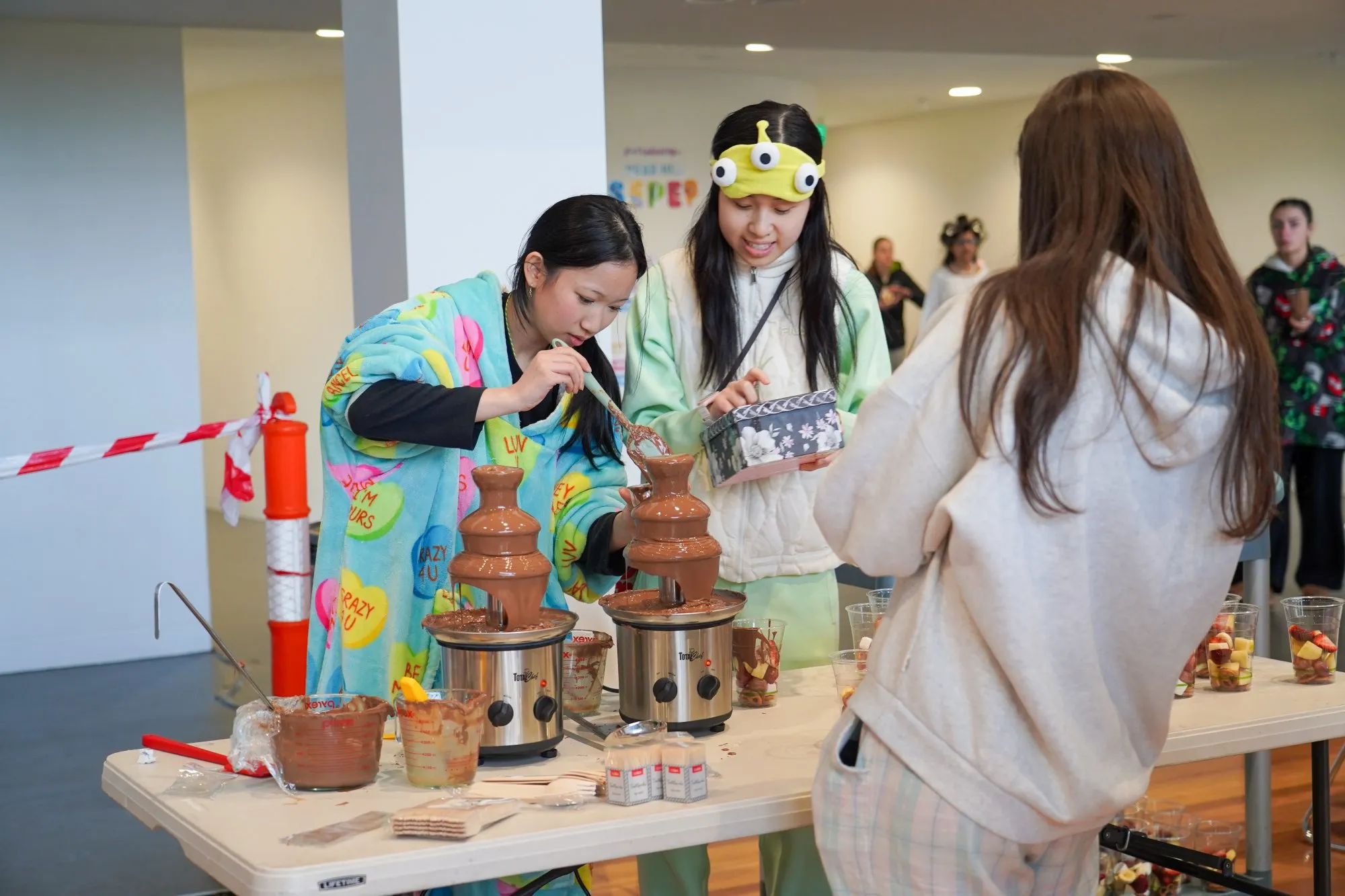 Three young women at a table with two chocolate fountains and cups of fruit for dipping indoors.