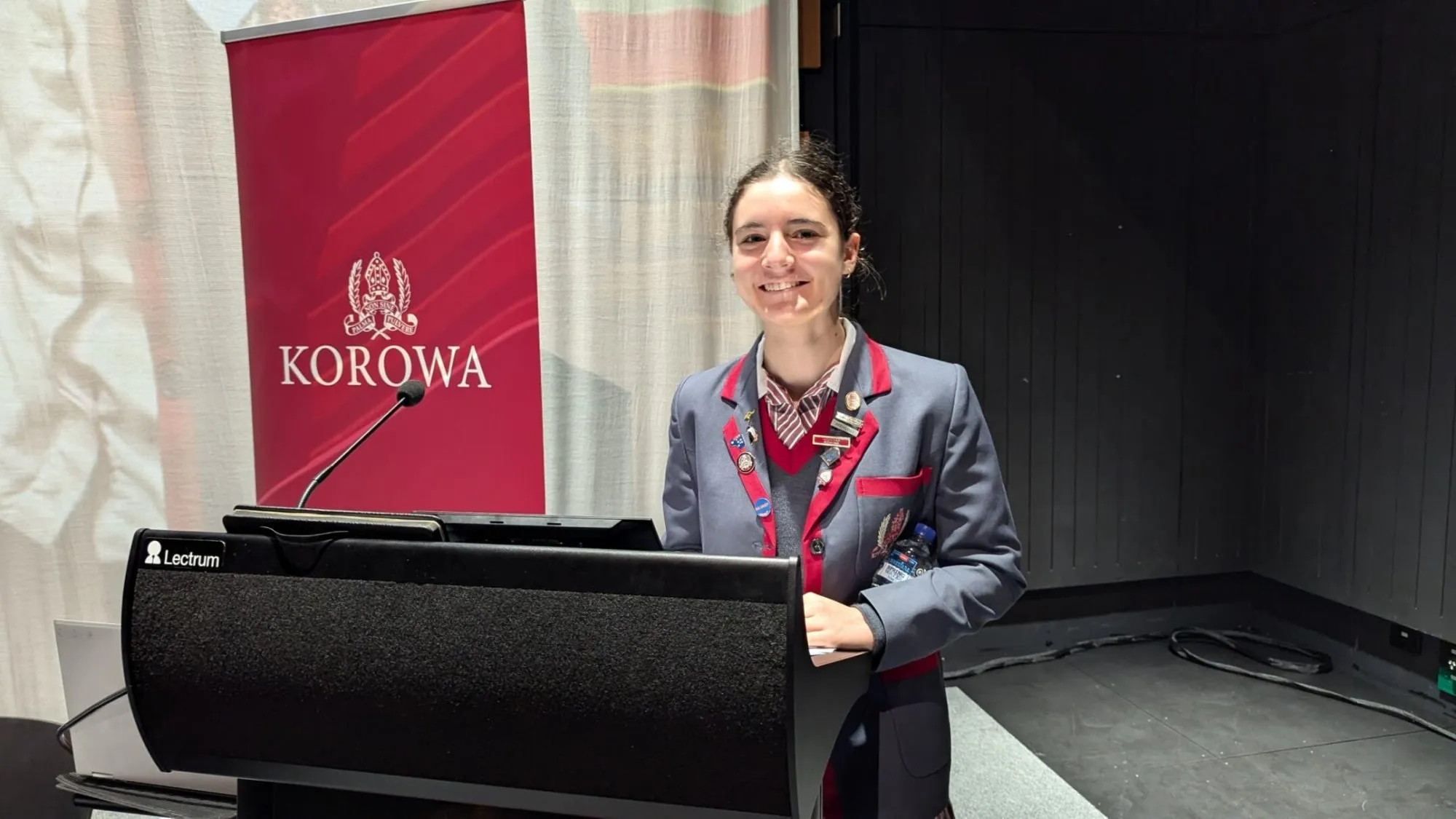 Smiling student in a gray blazer with red trim standing behind a lectern with a Korowa banner in the background.
