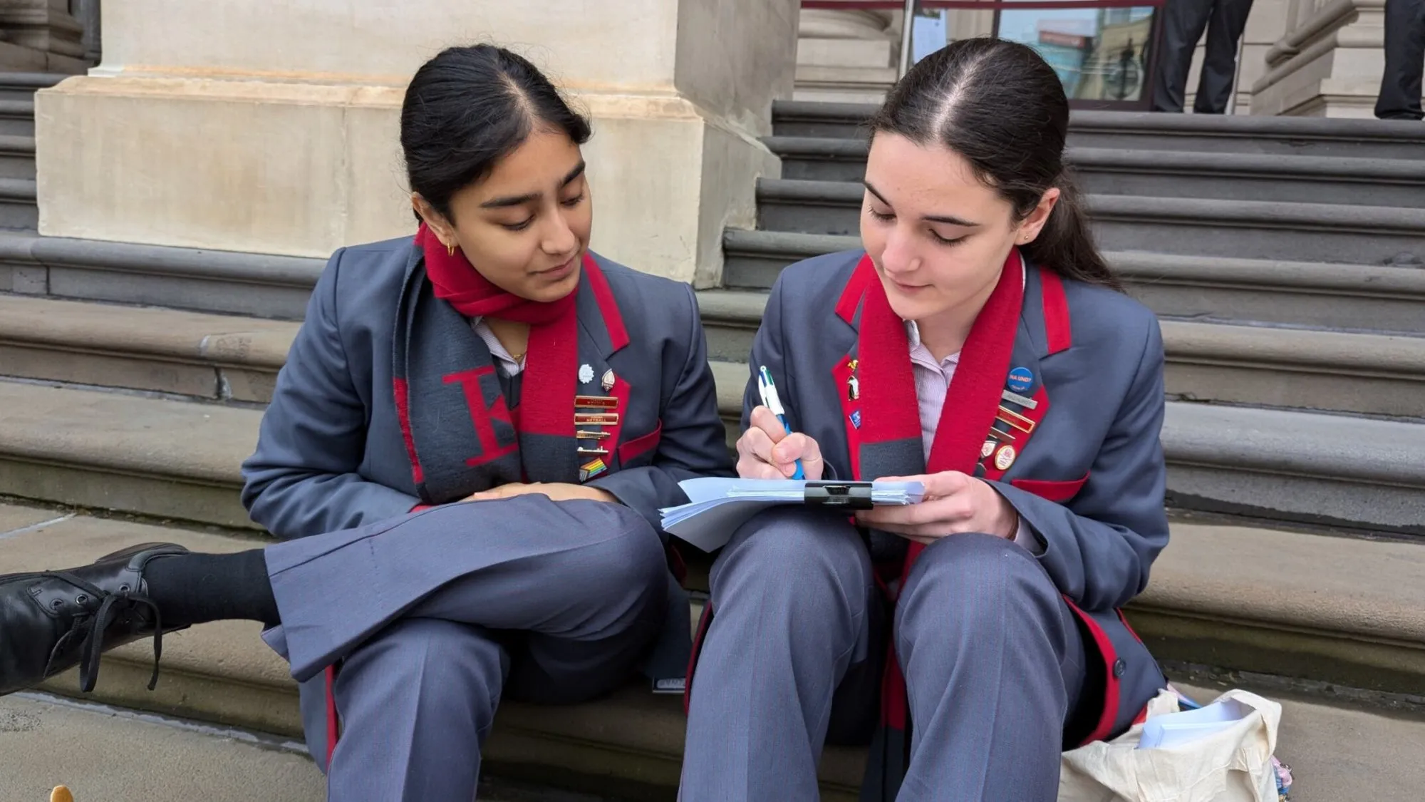 Two female students in gray uniforms with red trim and scarves sitting on stone steps, one writing in a notebook while the other watches.