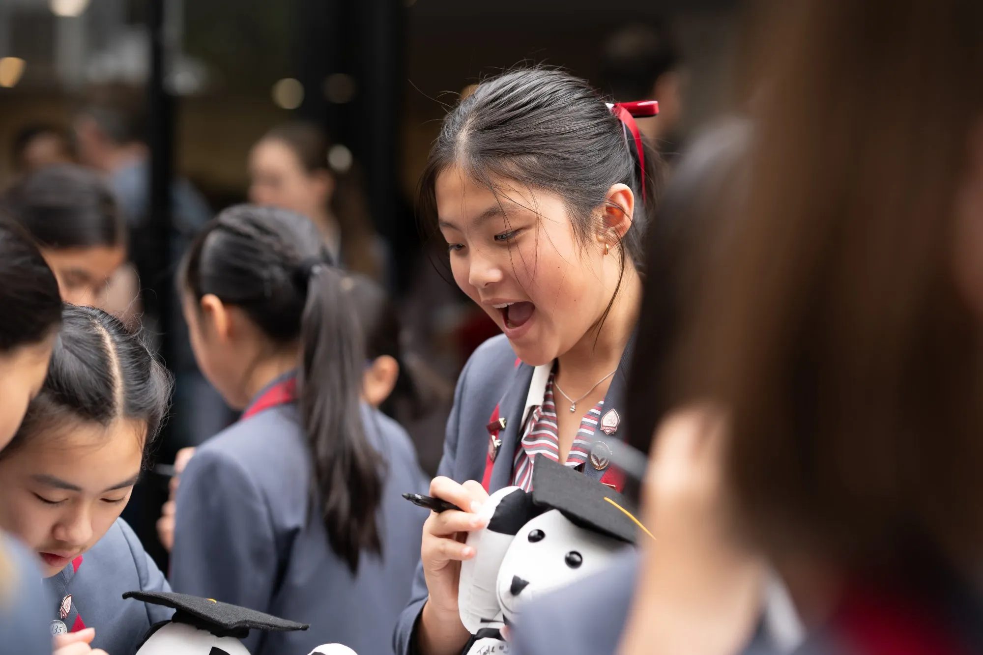 Excited schoolgirl in uniform holding a stuffed dog wearing a graduation cap, surrounded by other students.