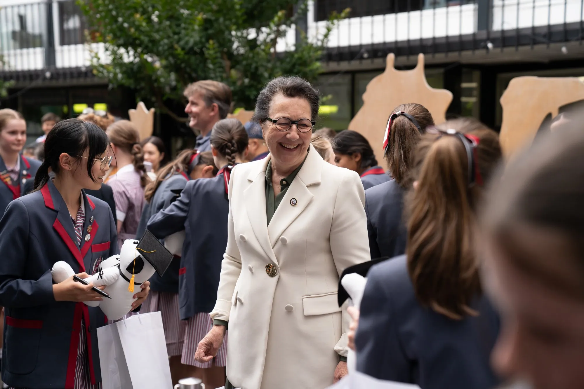 Smiling woman in white blazer interacts with students in school uniforms holding plush toys with graduation caps.