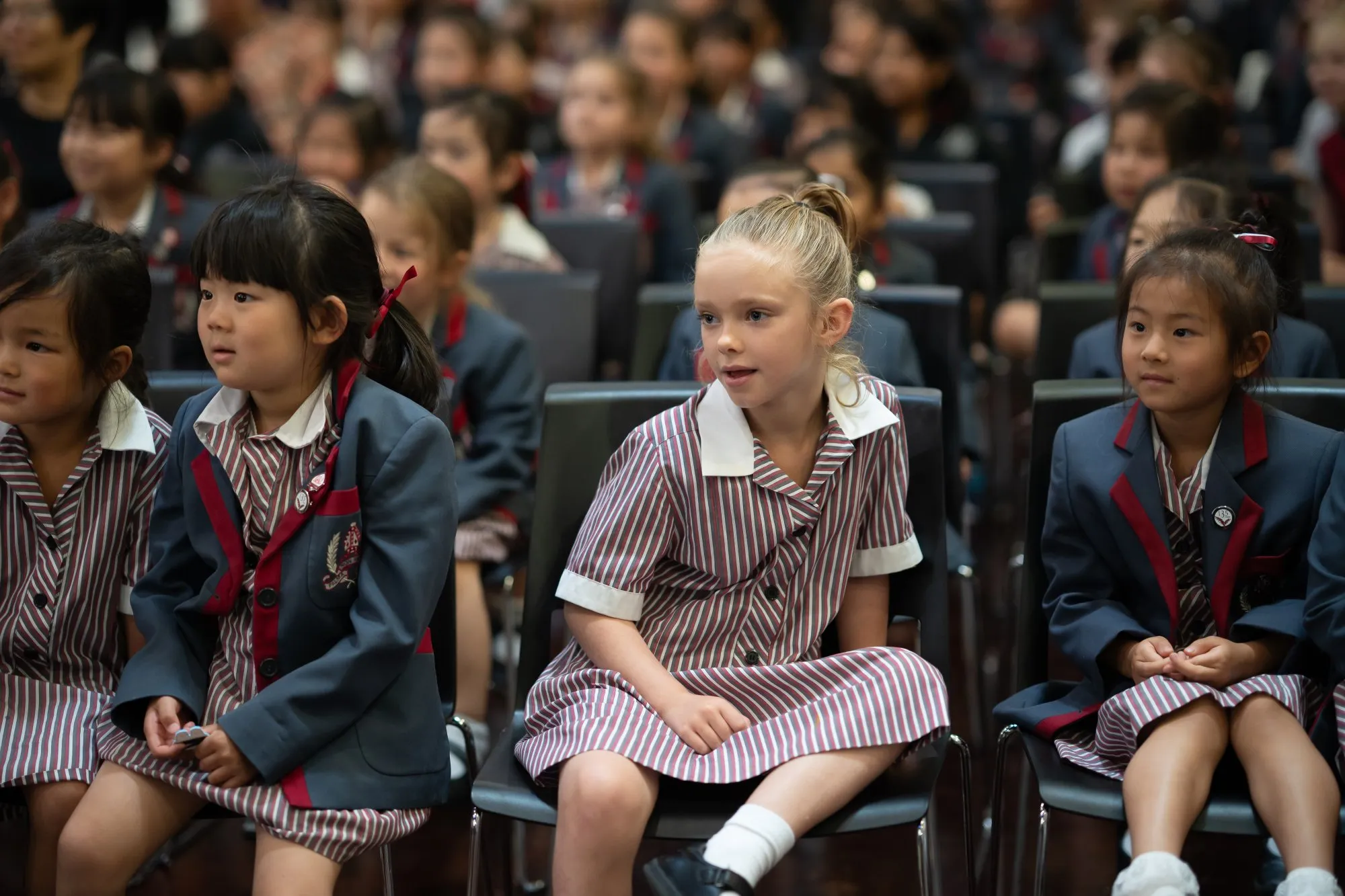 Young students in striped uniforms and navy blazers sitting attentively in chairs in a school assembly.