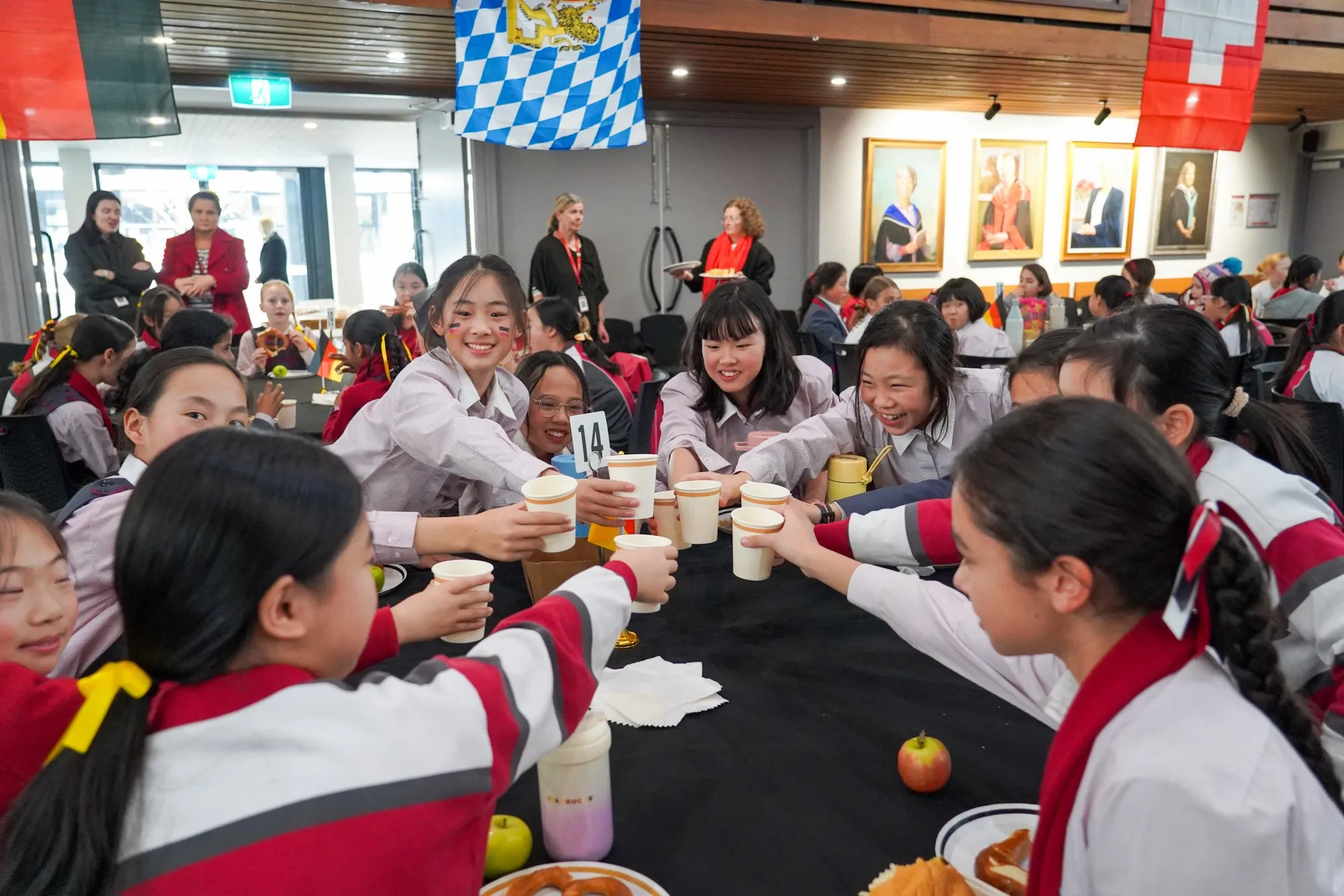 Group of schoolgirls in uniforms smiling and toasting with paper cups around a table at an indoor event with German and Swiss flags visible.