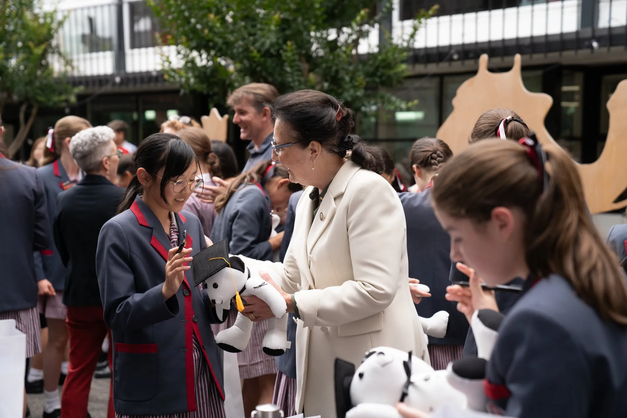 Smiling woman in white blazer handing a graduation-capped stuffed dog to a girl in a school uniform at an outdoor school event.