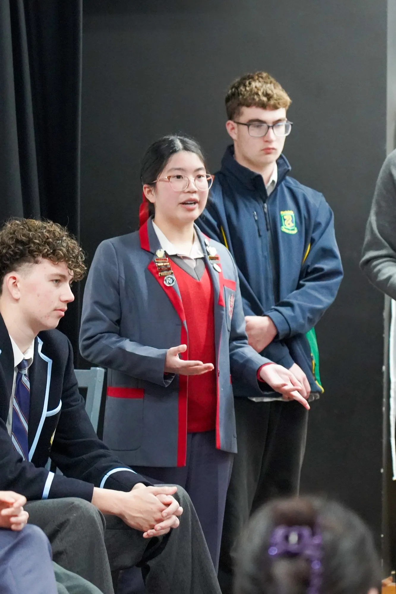 A young woman in a grey and red school uniform speaks, flanked by two young men in school uniforms standing and sitting against a dark background.