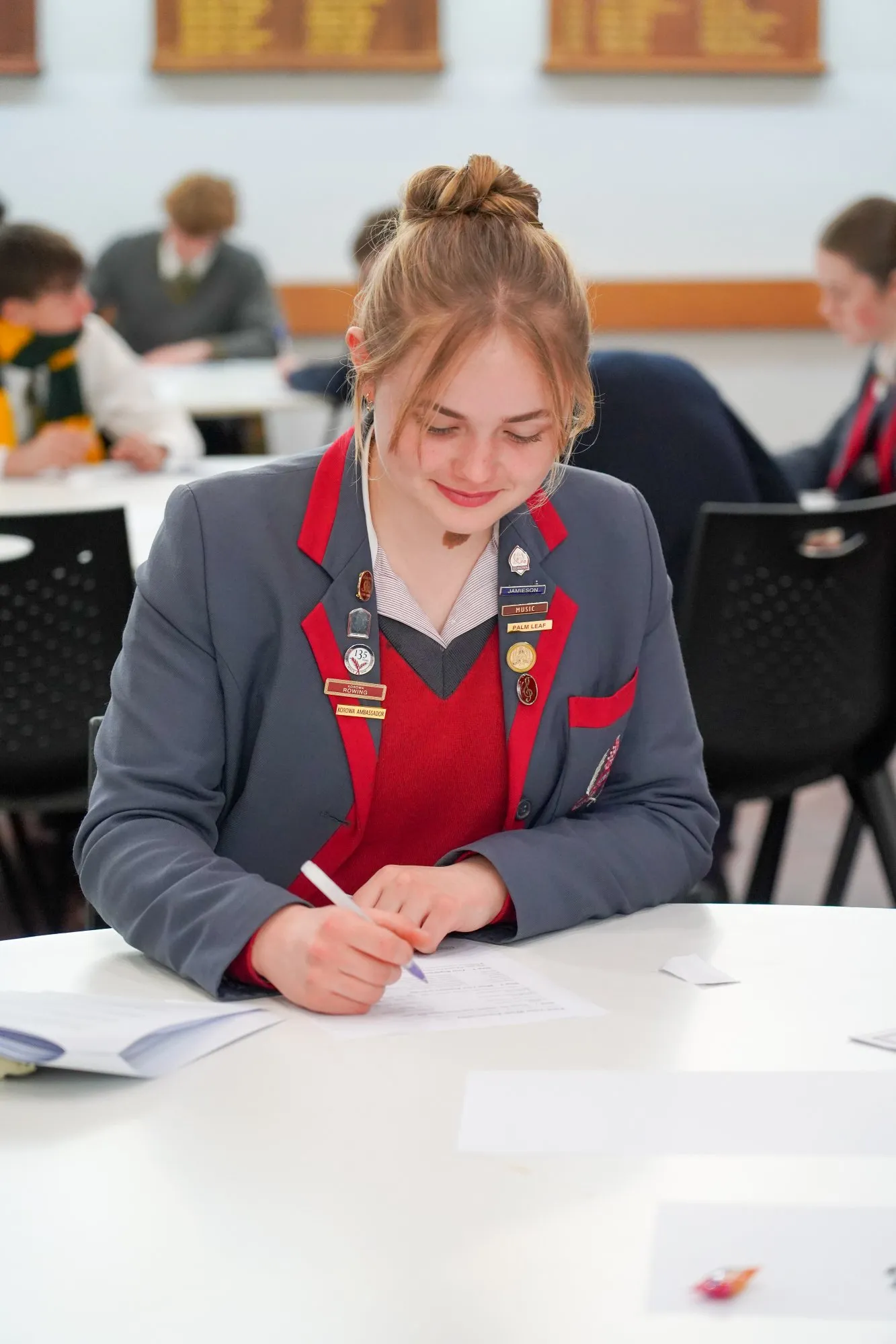 Student in a gray and red school uniform writing on paper at a round table in a classroom.