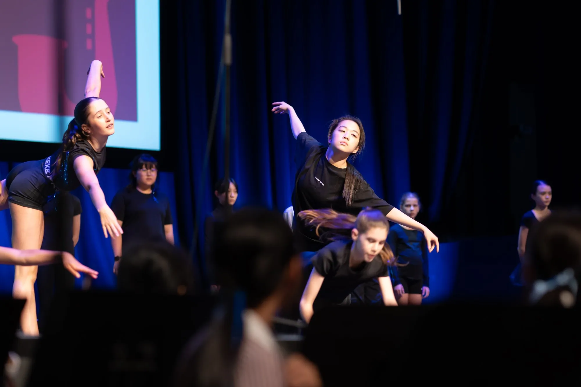 Young dancers in black outfits performing contemporary dance on stage with blue curtains in the background.