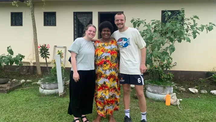 Three people standing on green grass in front of a beige building, smiling with their arms around each other; the person in the middle wears a colorful dress.