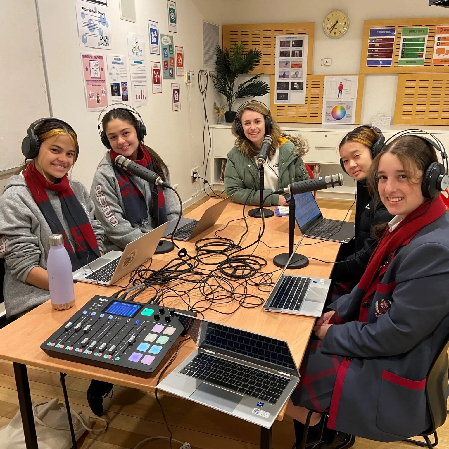 Five young women wearing headphones sitting around a table with laptops and microphones in a podcast recording setup.