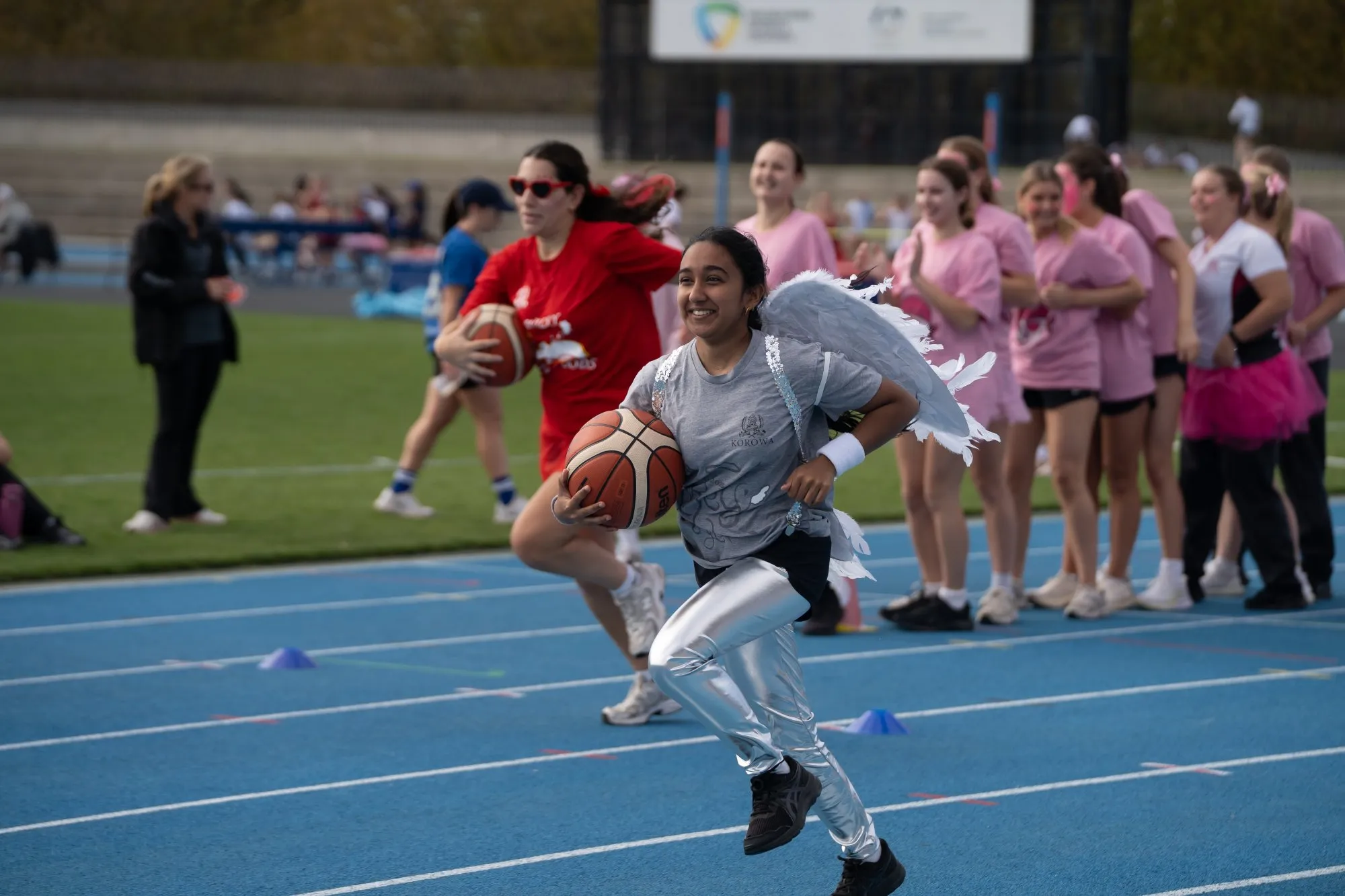 Smiling girl wearing angel wings and shiny silver pants running with a basketball on a blue track, pursued by another player in red, with a group of girls in pink shirts cheering in the background.