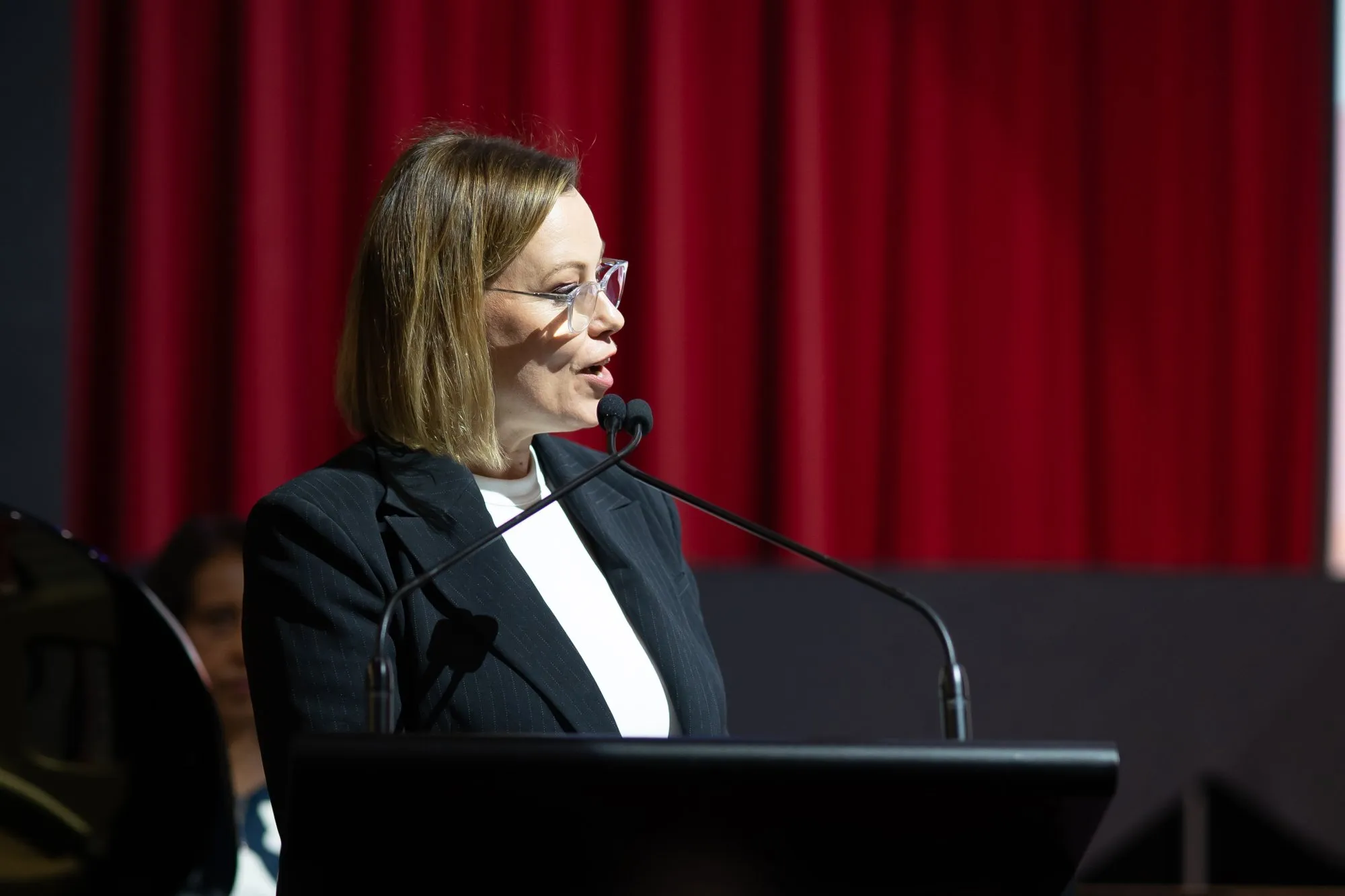 Woman in glasses speaking at a podium with two microphones against a red curtain background.