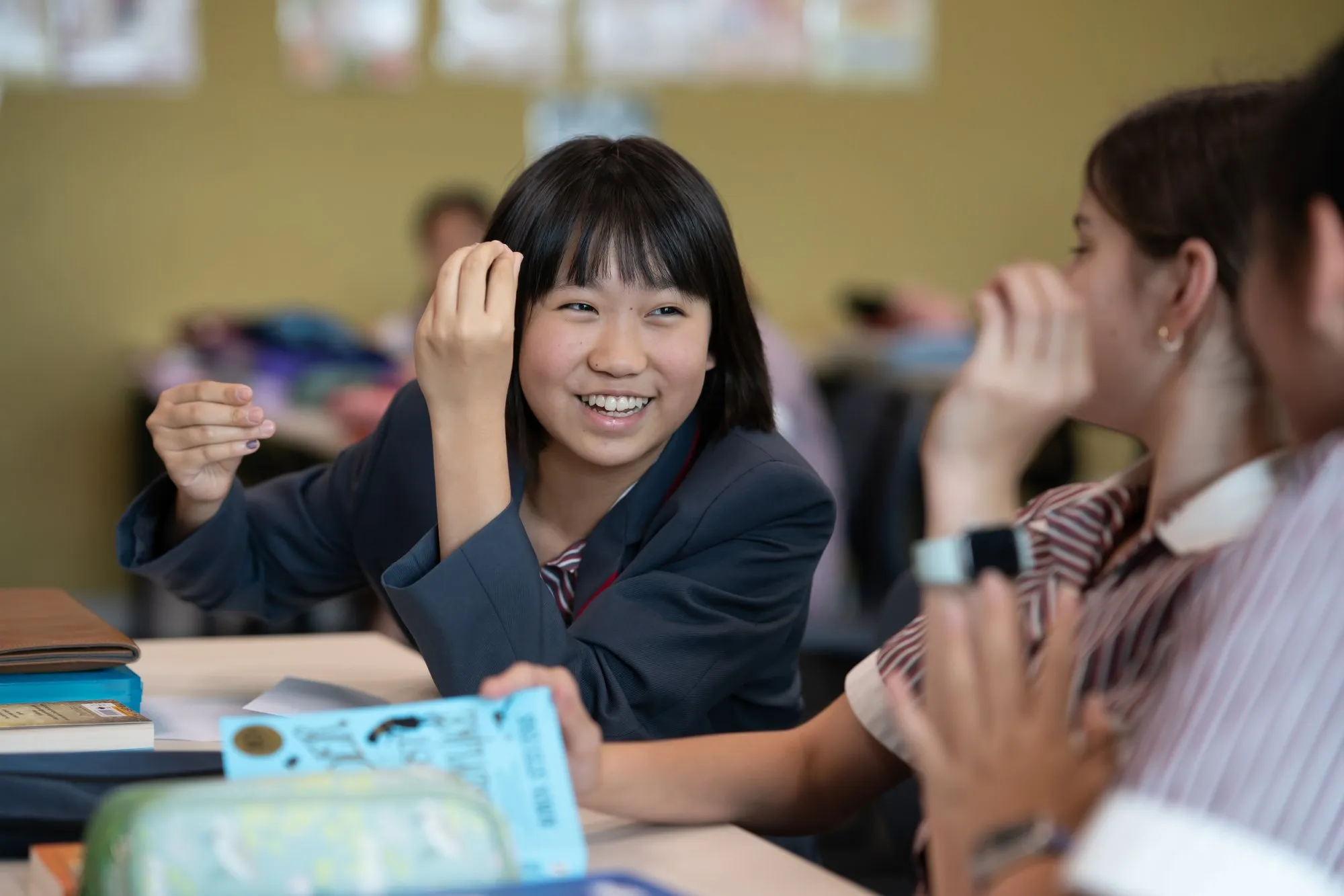 Smiling schoolgirl in uniform engaging in conversation with classmates at a classroom desk.