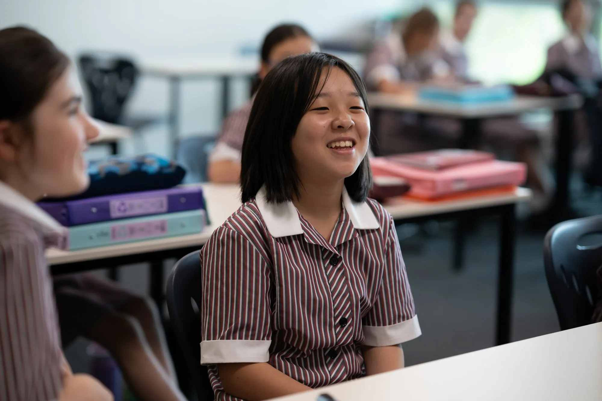 Smiling schoolgirl in a striped uniform sitting at a classroom desk with other students in the background.