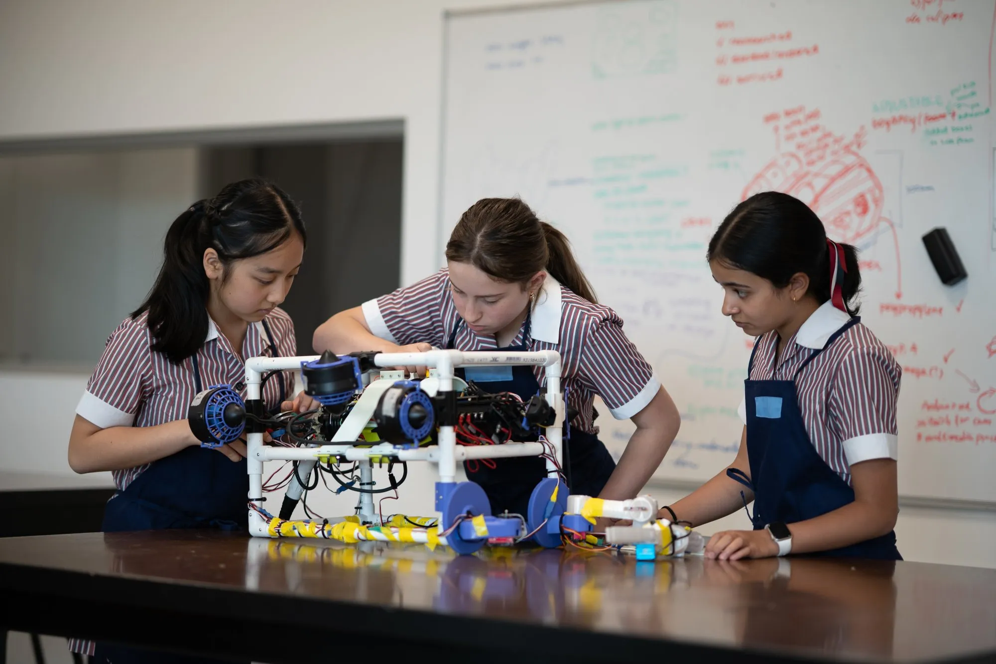 Three girls in striped school uniforms and aprons working together on assembling a robotic device on a table.