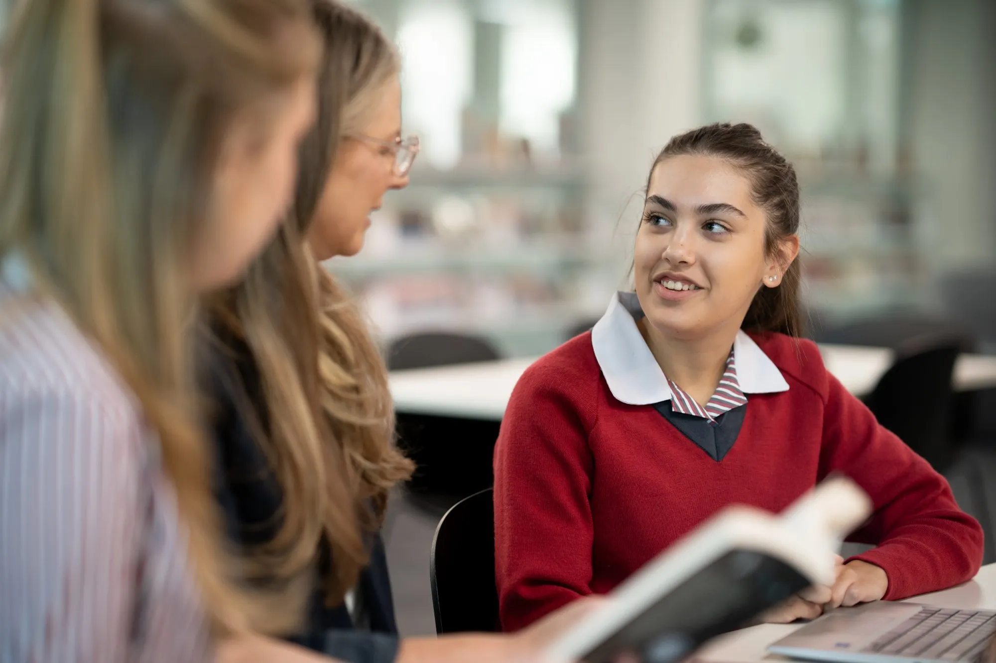 A student in a red sweater smiling and interacting with two adults in a classroom setting.