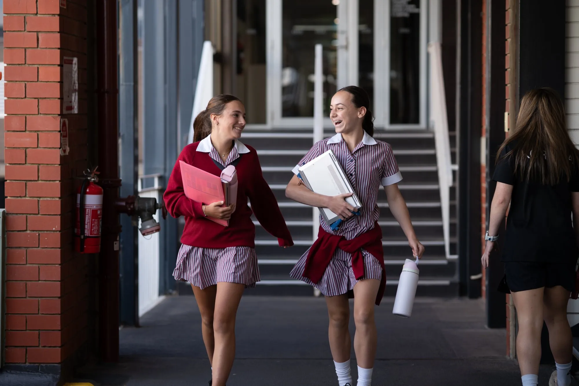 Two female students in striped school uniforms walking in a hallway, smiling and holding books and a water bottle.