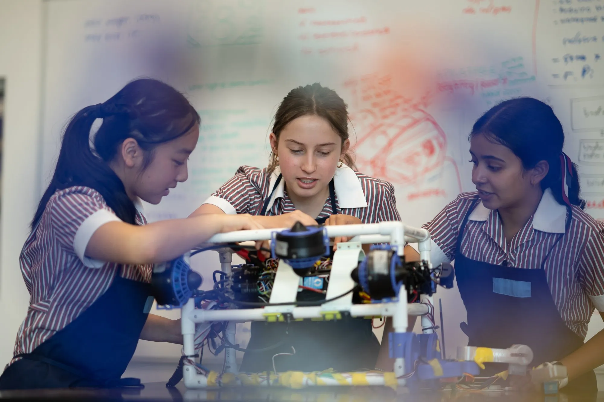 Three female students in striped uniforms and aprons working together on assembling a robotic device in a classroom.