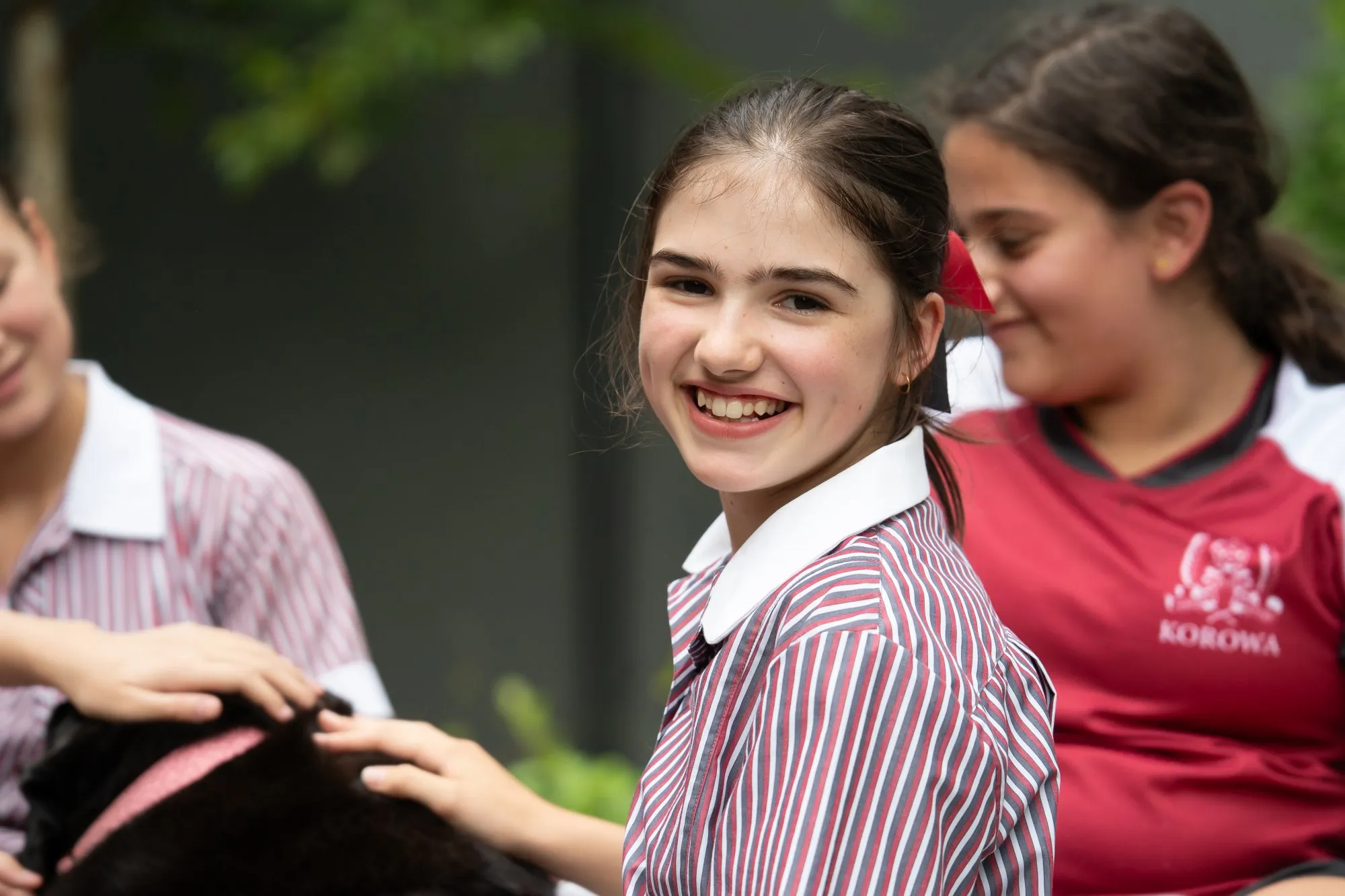 Smiling girl in a striped school uniform petting a black dog with other girls nearby.