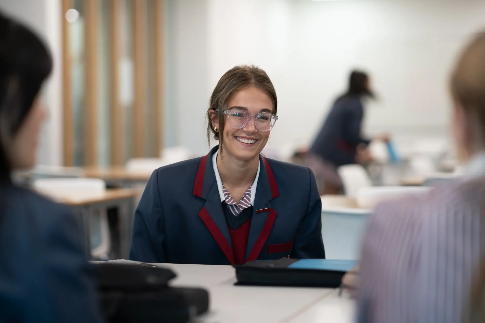 Smiling teenage student wearing glasses and a school uniform with a striped tie sitting at a table in a classroom.