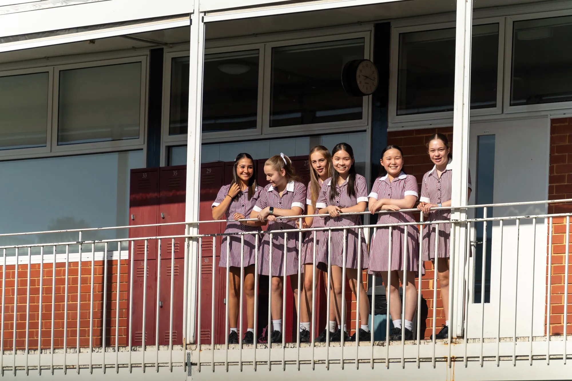 Six schoolgirls in striped uniforms smiling and standing on an outdoor balcony by red lockers and a clock.