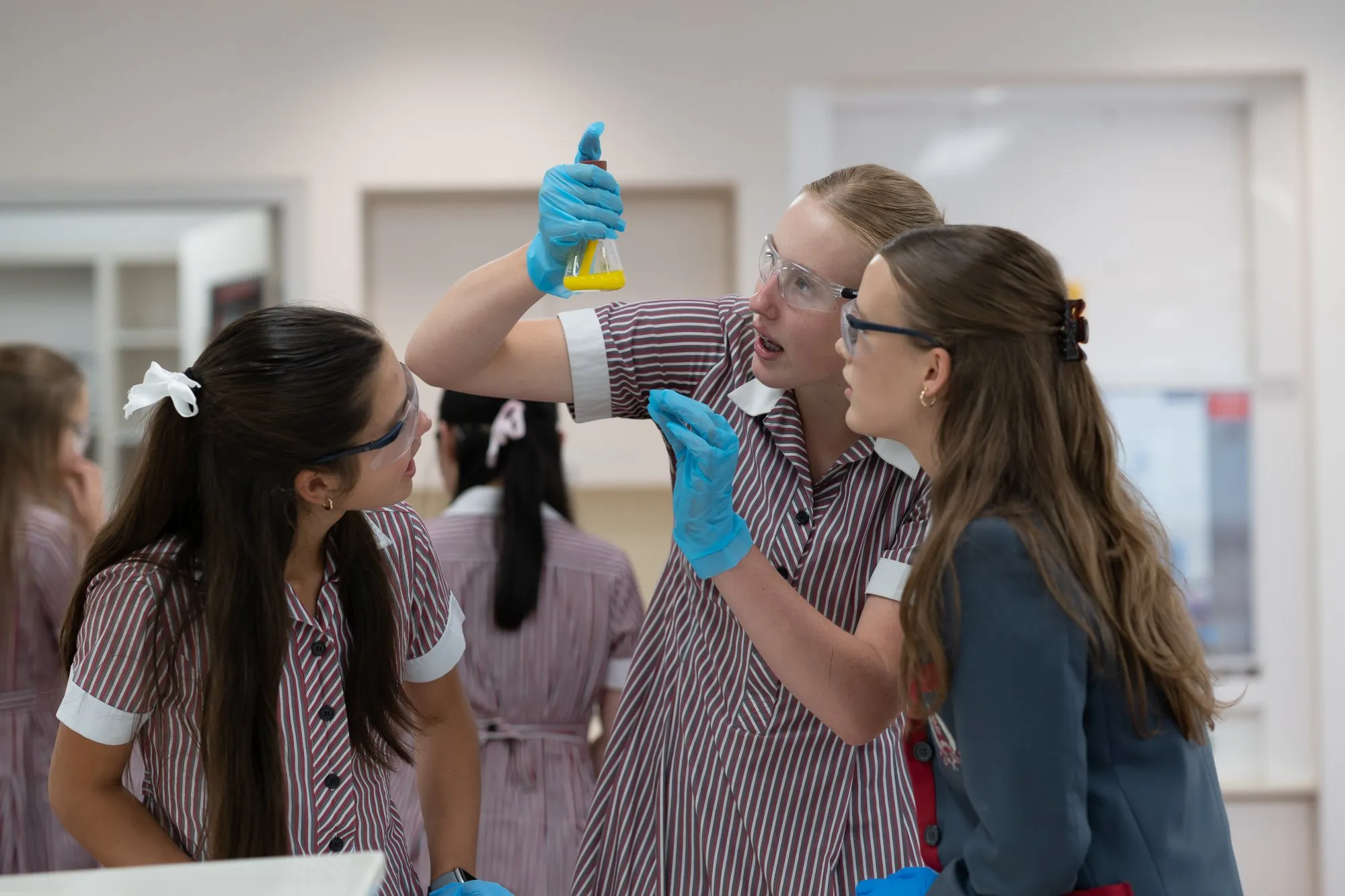 Three students wearing safety goggles and gloves closely examine a yellow liquid in a conical flask in a science lab.